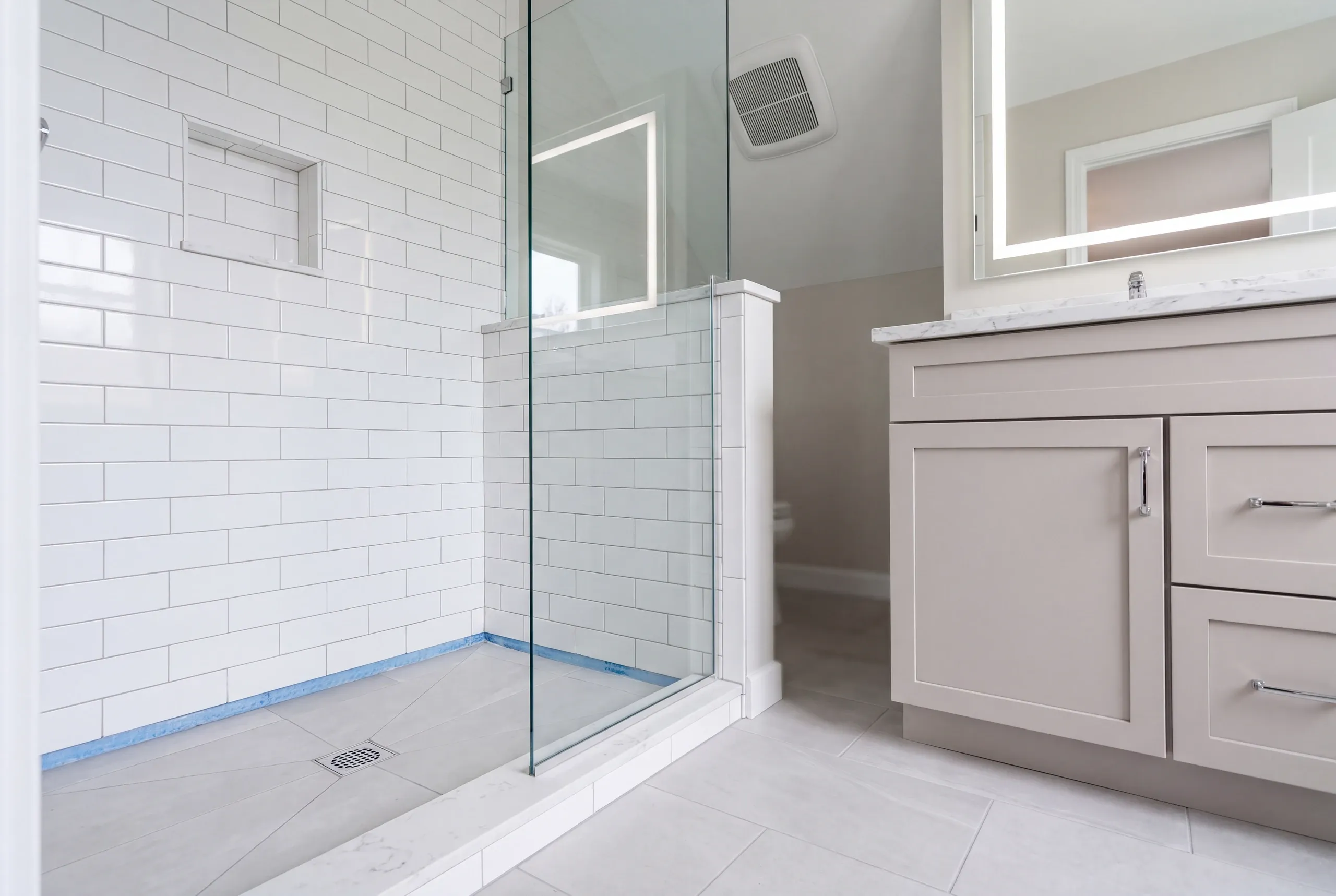 Bathroom renovation contractor Augusta Maine: nearly finished walk-in shower with a floor drain, glass panel and blue tape along the tile base