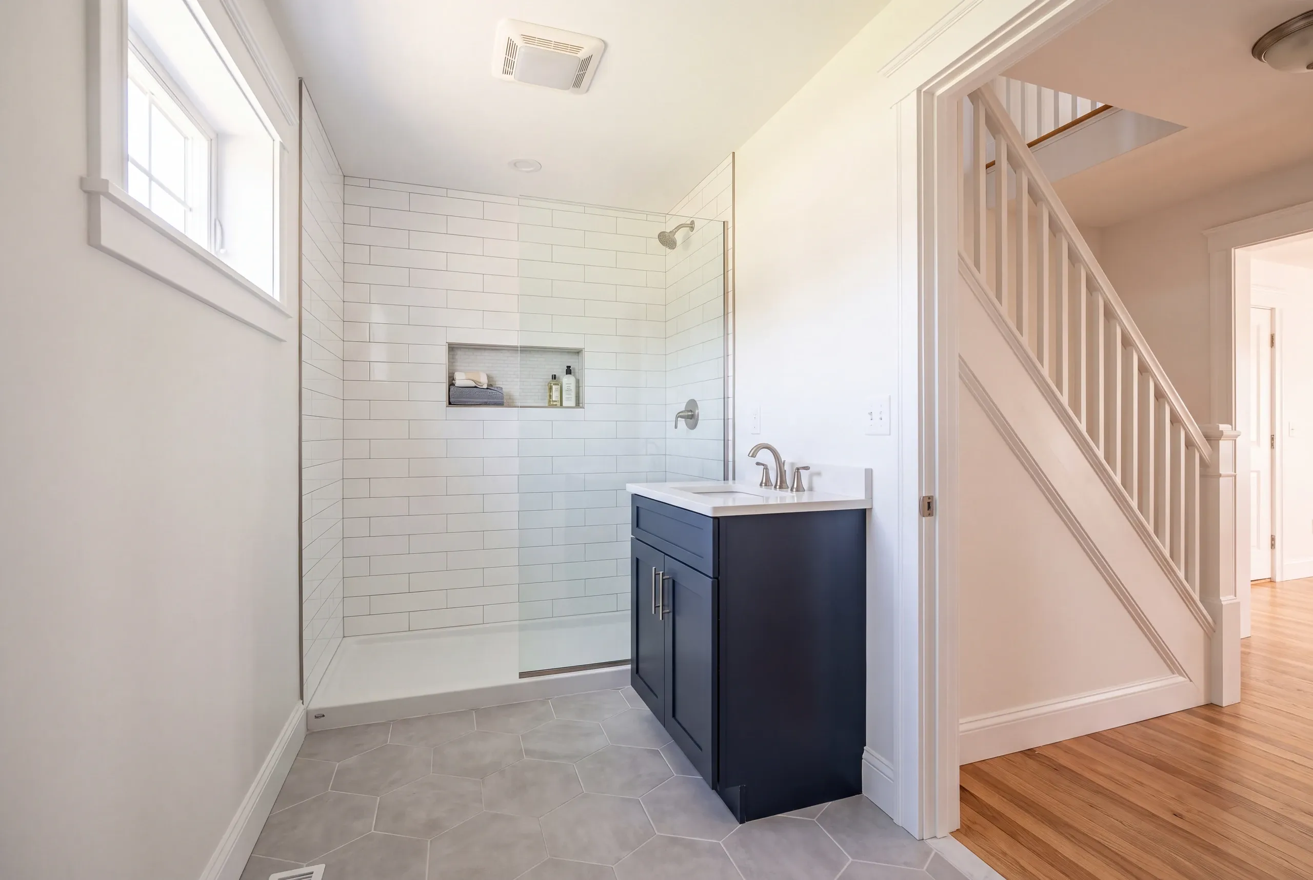 Bathroom remodeler Augusta Maine: full bathroom with a subway tile shower, navy vanity and hex floor, staircase seen through the door
