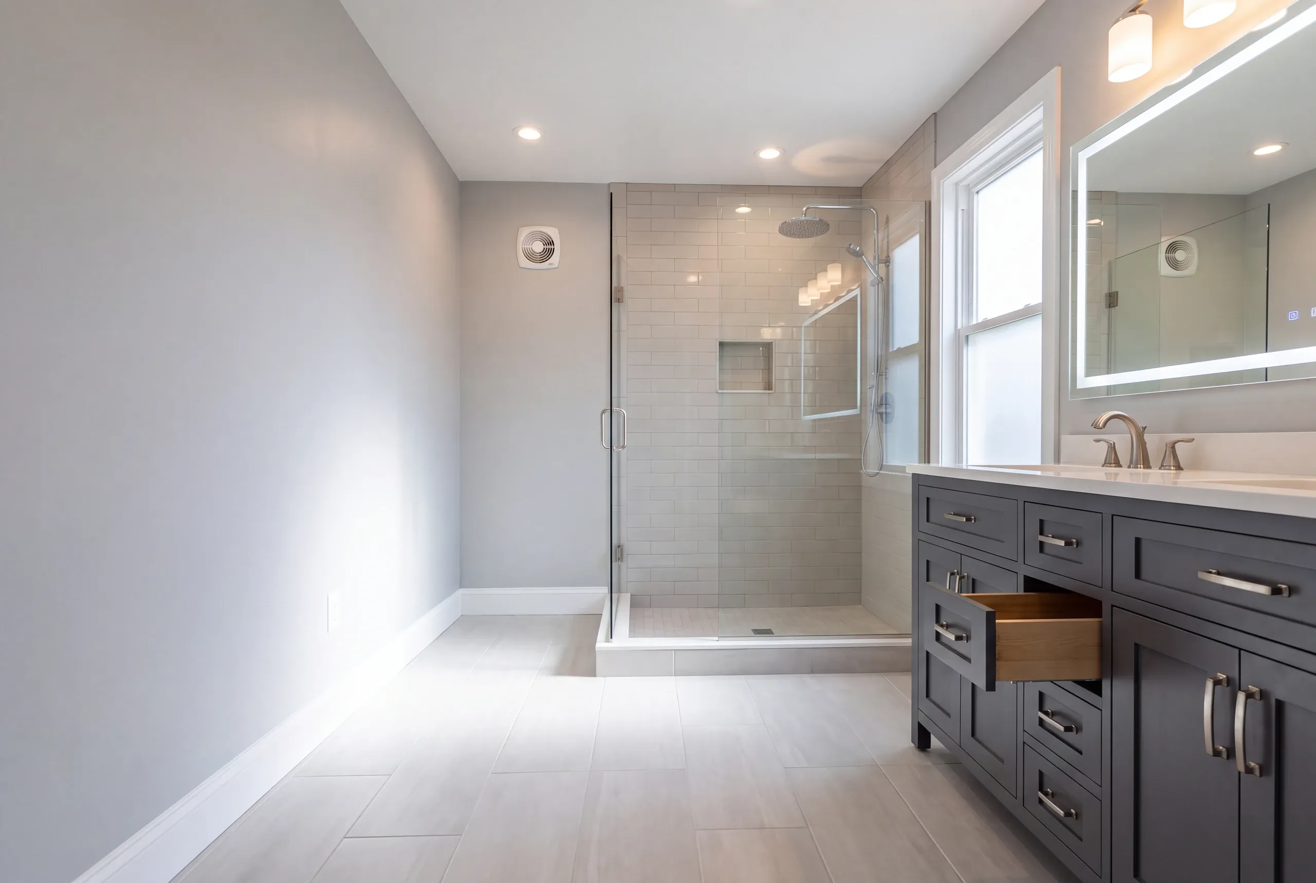 Bathroom remodeler Augusta Maine: long gray bathroom with a navy double vanity, open drawer and gray subway tile glass shower