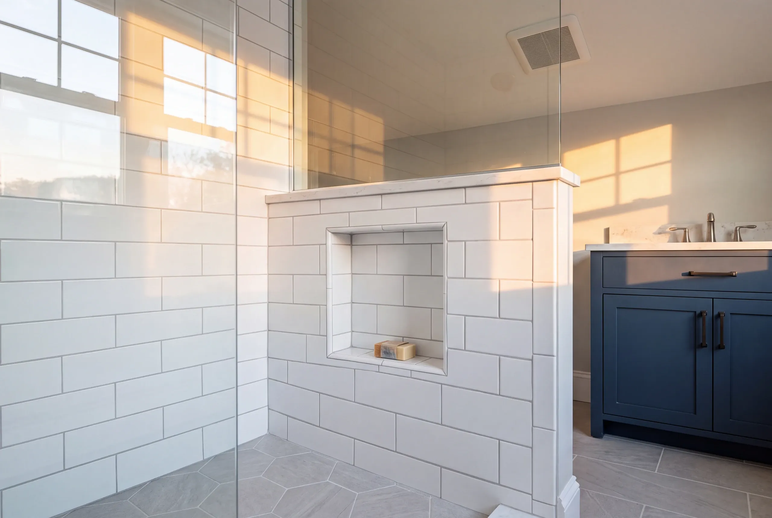 Bathroom renovation contractor Bath Maine: tiled shower half wall with a soap niche and glass panel beside a navy vanity and hex floor