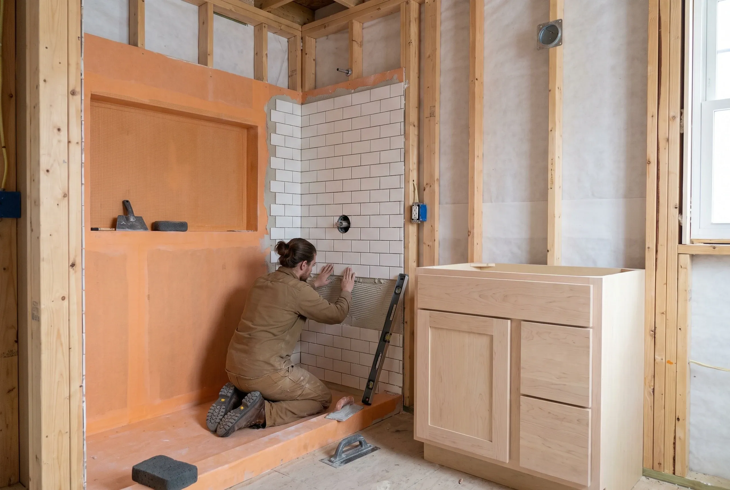 Bathroom remodeling contractor Belfast Maine: tile setter pressing subway tile onto a shower wall beside an orange membrane niche and vanity