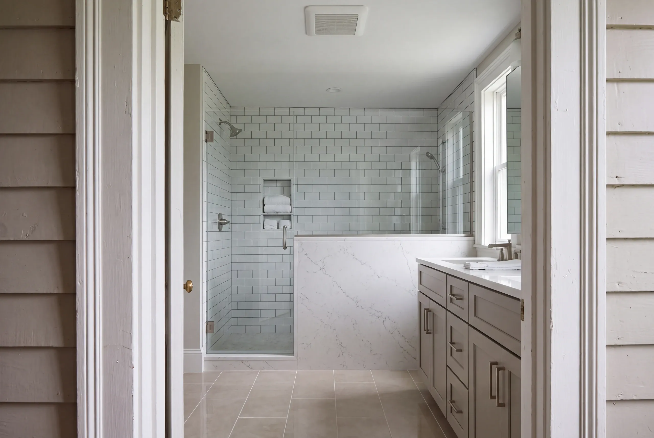 Bathroom remodeler Damariscotta Maine: doorway view of a subway tile shower with a stone-capped half wall and gray shaker vanity
