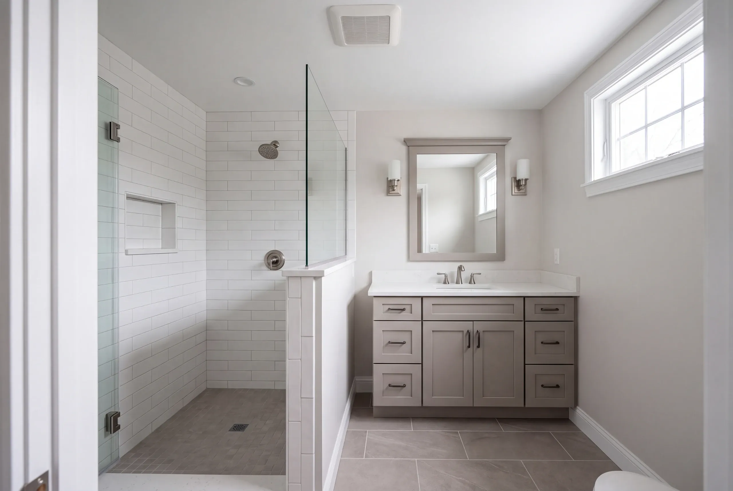 Bathroom remodeling contractor Fairfield Maine: subway tile shower with a glass door and half-wall panel beside a gray vanity and mirror