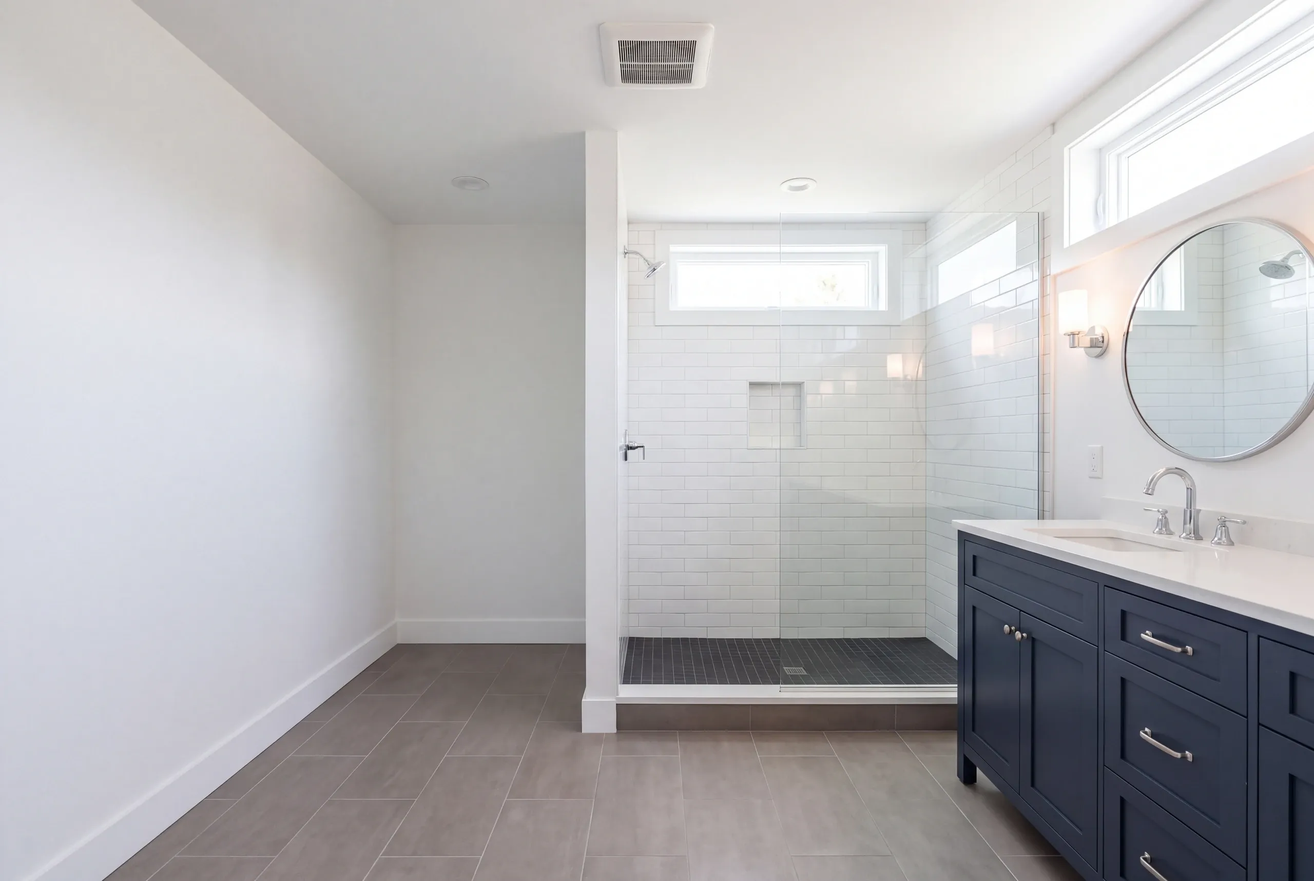 Bathroom remodeler Lincolnville Maine: bathroom with a dark mosaic shower floor under transom windows, navy vanity and round mirror