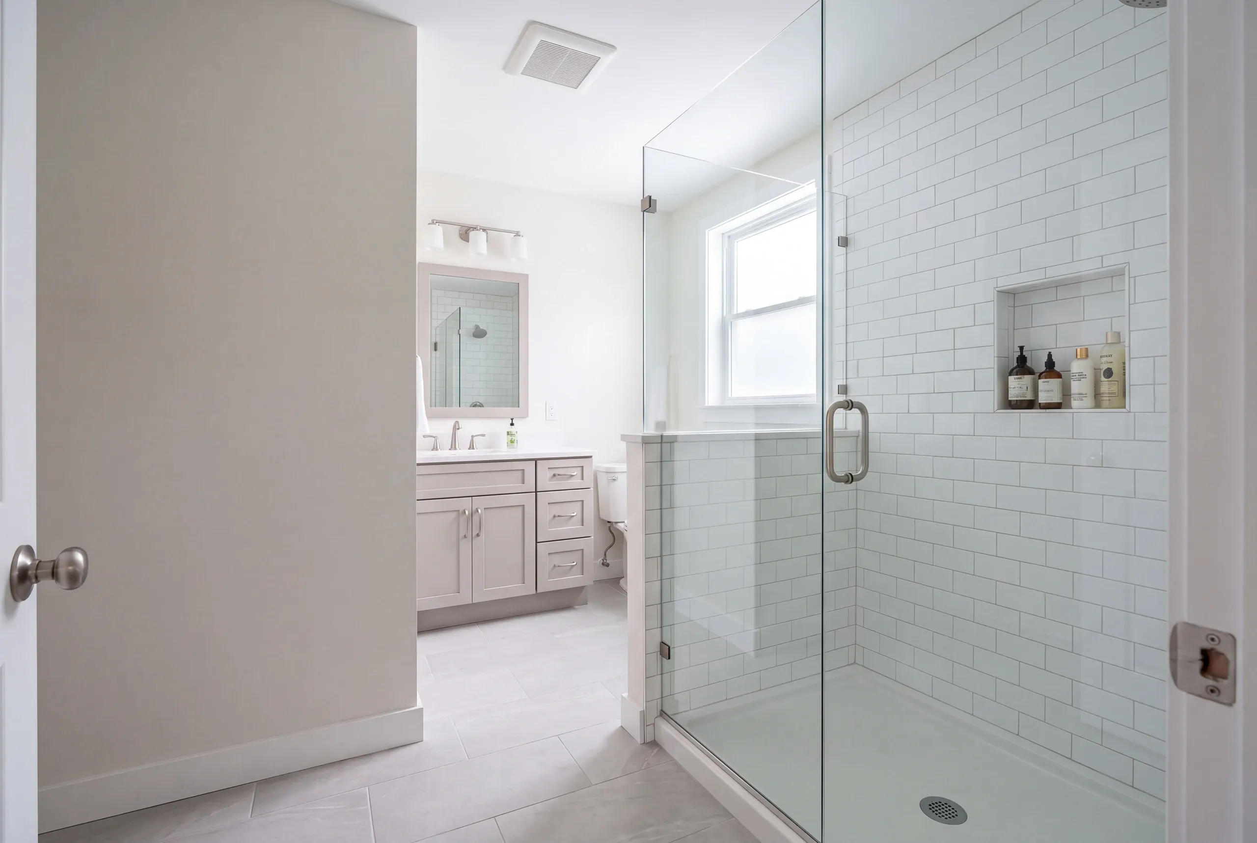Bathroom renovation contractor Pittsfield Maine: doorway view of a glass corner shower with subway tile and a gray vanity beyond