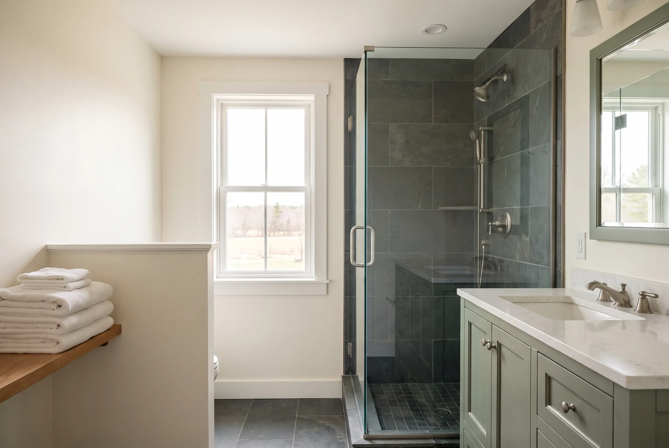 Bathroom remodeling contractor Pittston Maine: slate tile shower with glass door beside a sage vanity and framed mirror