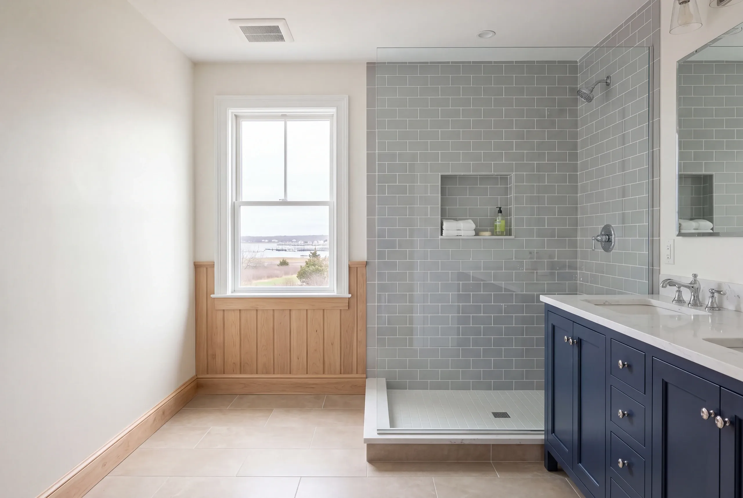 Bathroom remodeler Rockland Maine: bathroom with a gray subway tile shower, navy double vanity and wood wainscot by a water view
