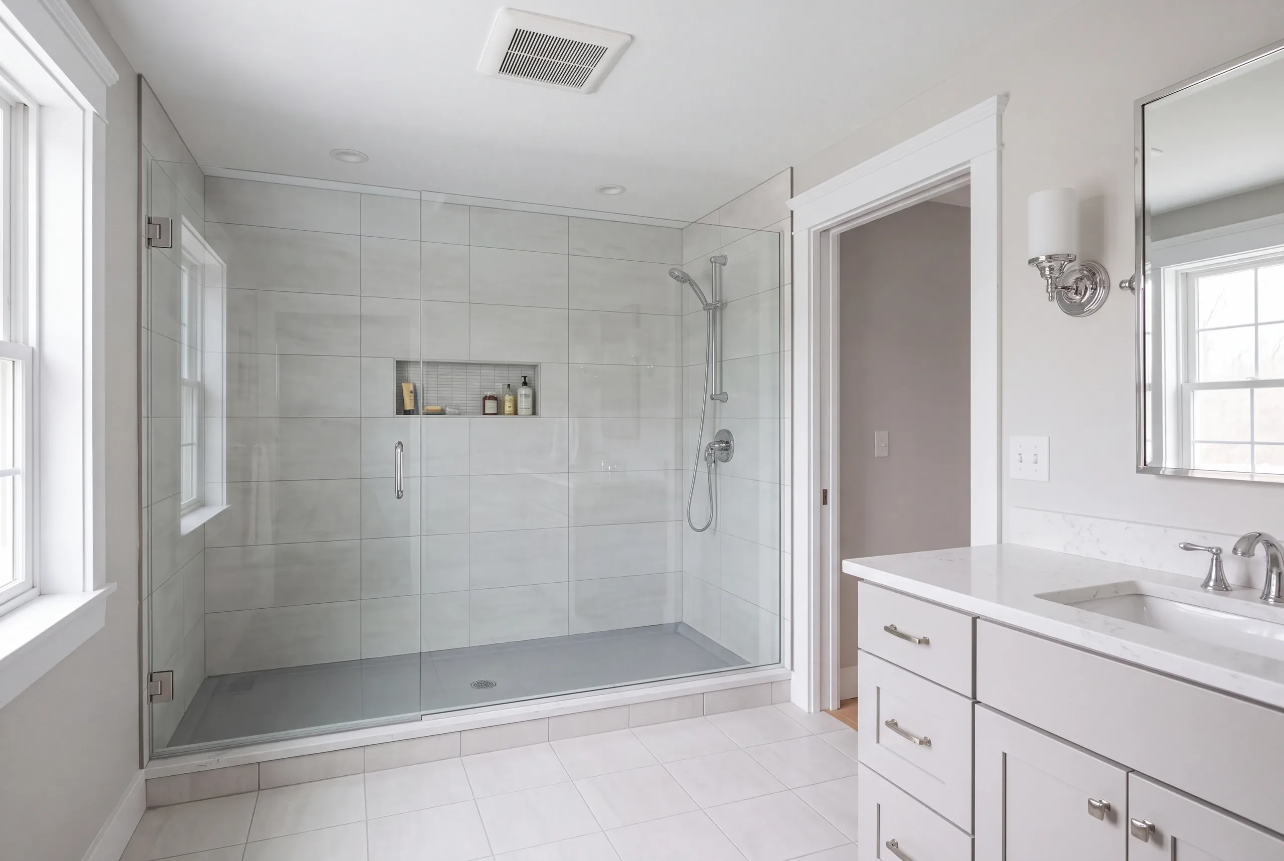 Bathroom remodeling in Searsmont, ME: light gray large tile shower with a glass door and niche beside a white vanity with a stone-look top