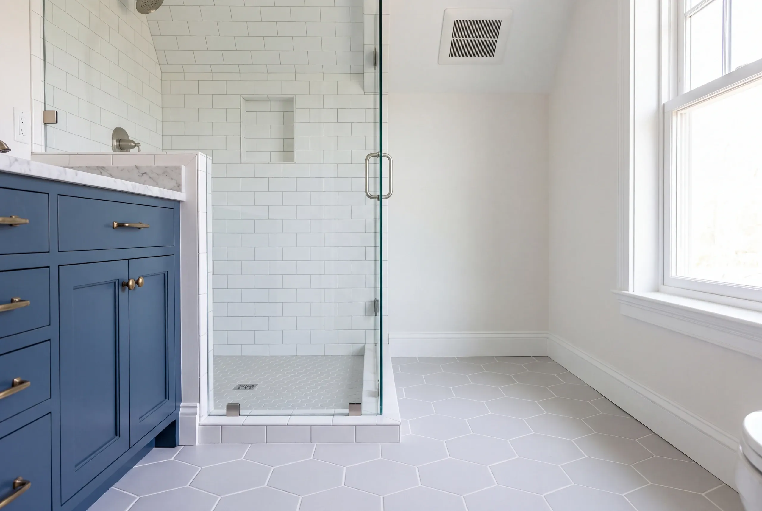 Bathroom renovation contractor Thomaston Maine: blue vanity with brass pulls beside a glass corner shower and large gray hex floor tile