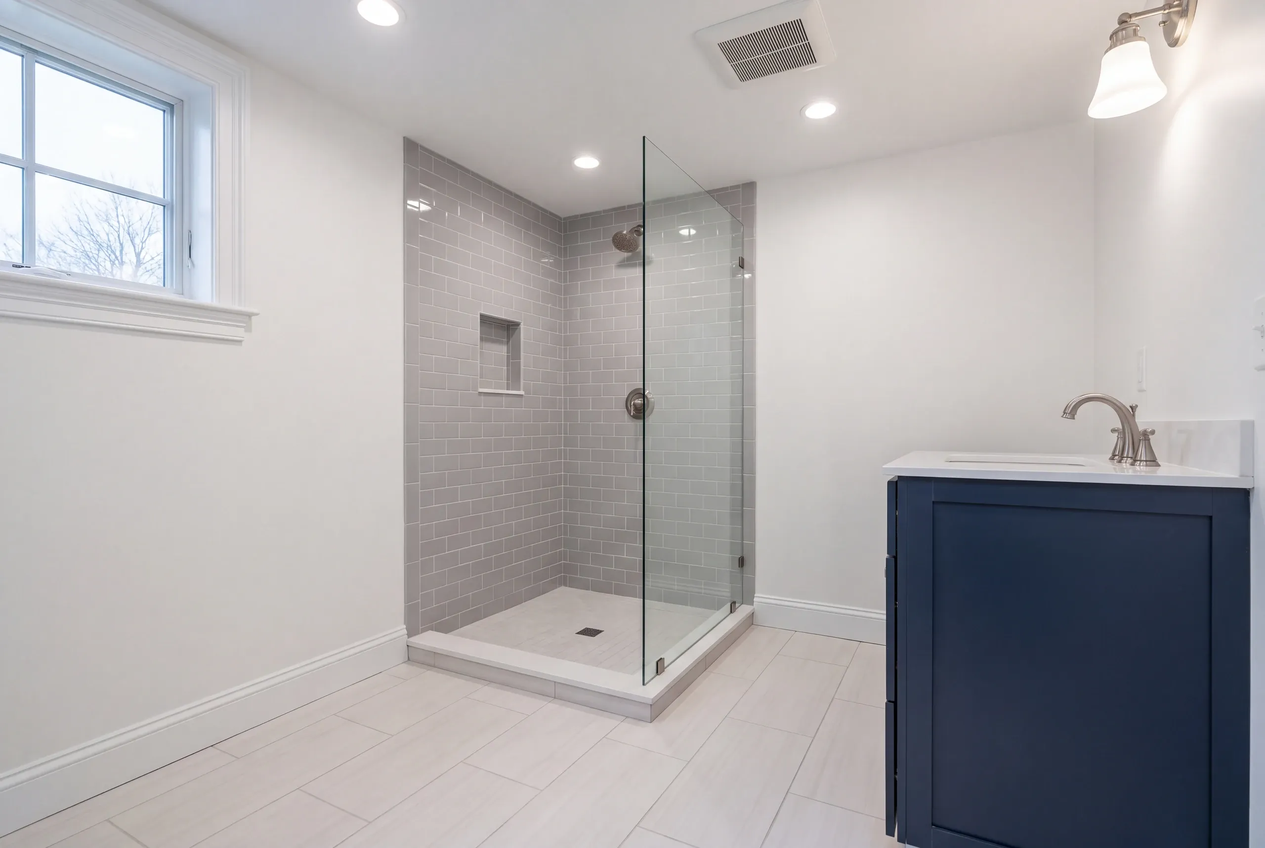 Bathroom renovation contractor Thomaston Maine: gray subway tile corner shower with a glass panel beside a navy vanity and window