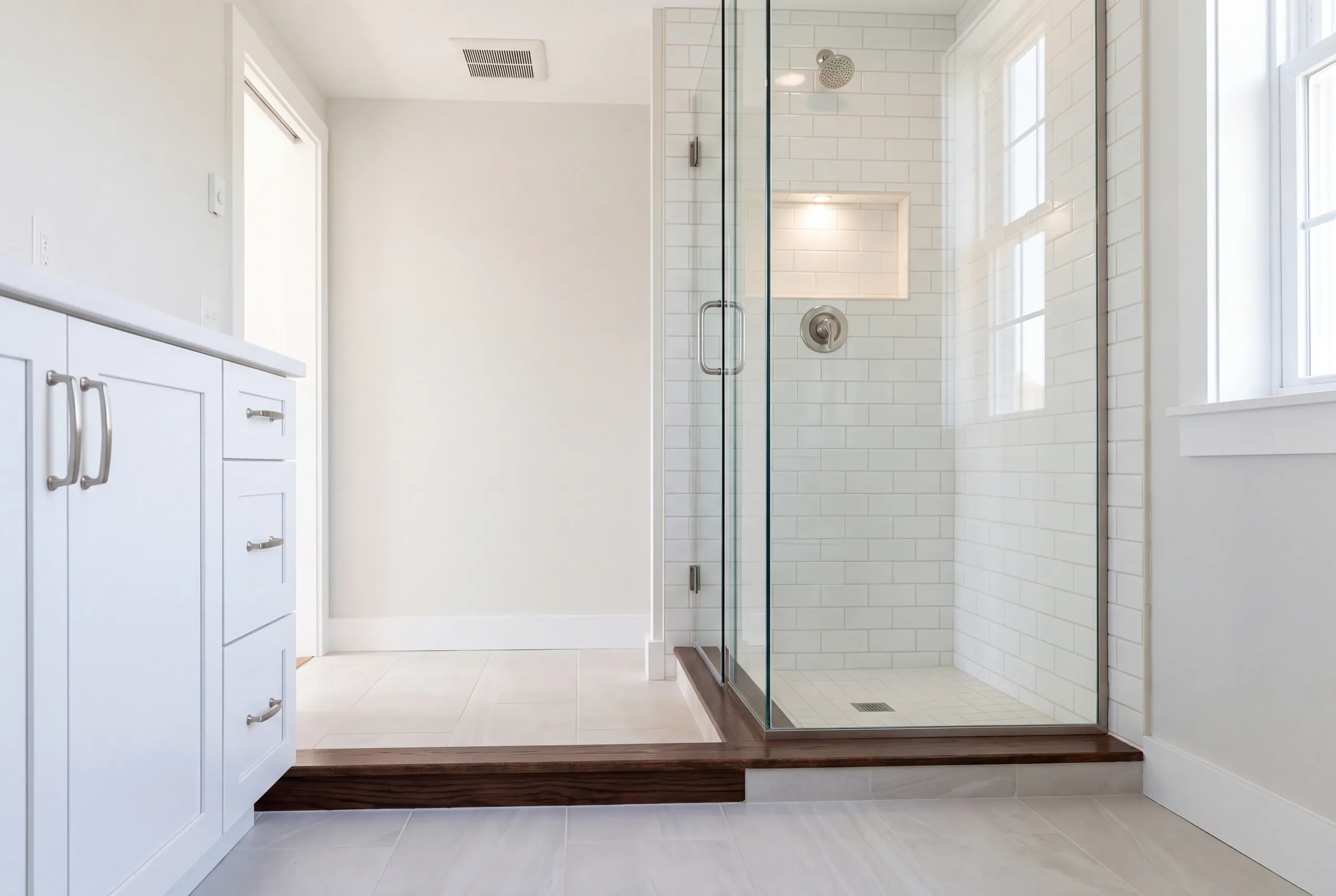Bathroom renovation contractor Waldoboro Maine: walnut threshold strip at a raised floor beside a glass subway tile shower and white vanity