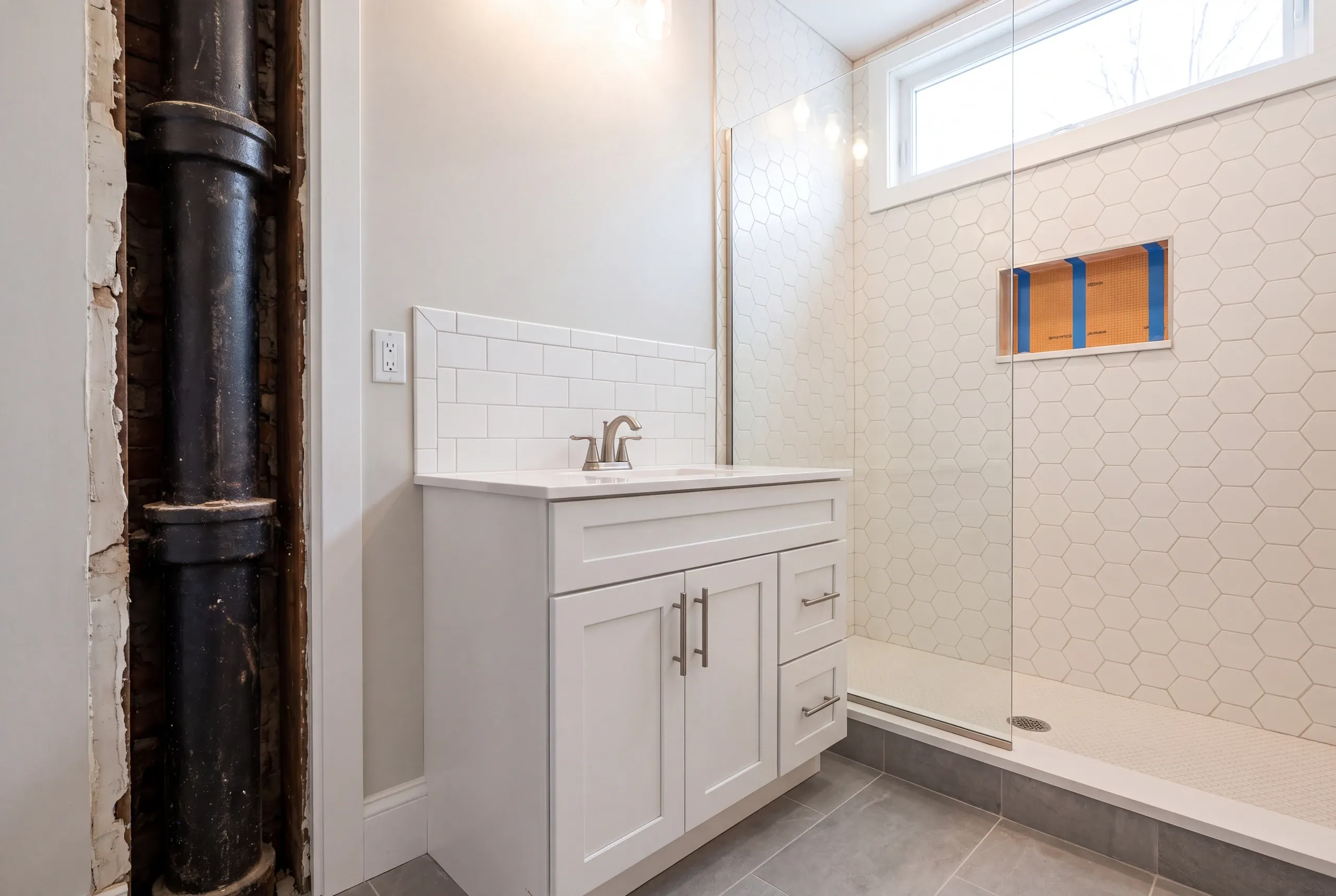 Bathroom renovation contractor Waterville Maine: open wall showing an old black cast iron stack beside a white vanity and hex tile shower