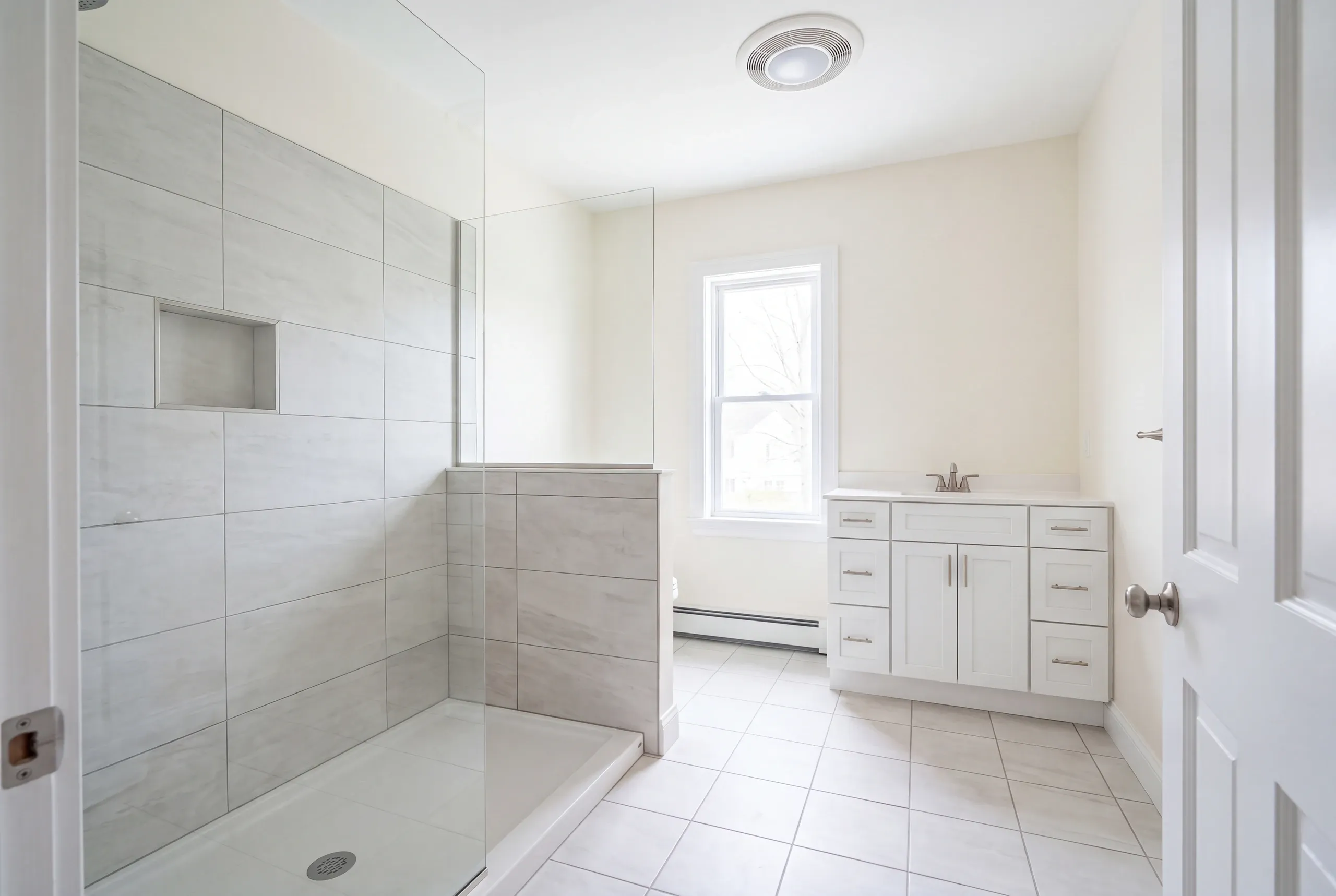 Bathroom remodeler Waterville Maine: light gray tile shower on a white base behind glass with a white vanity beyond