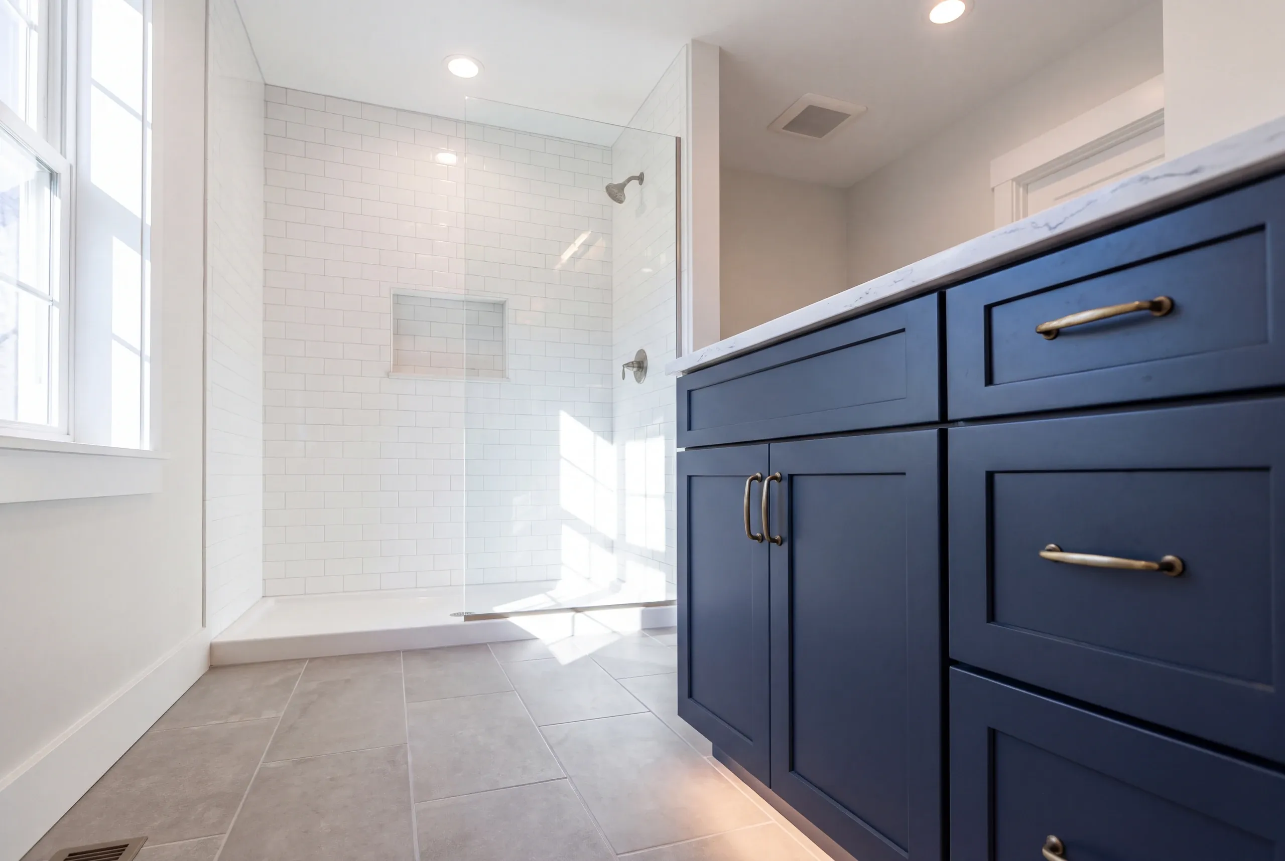 Bathroom renovation contractor Windsor Maine: navy vanity with brass pulls beside a walk-in subway tile shower and gray floor tile