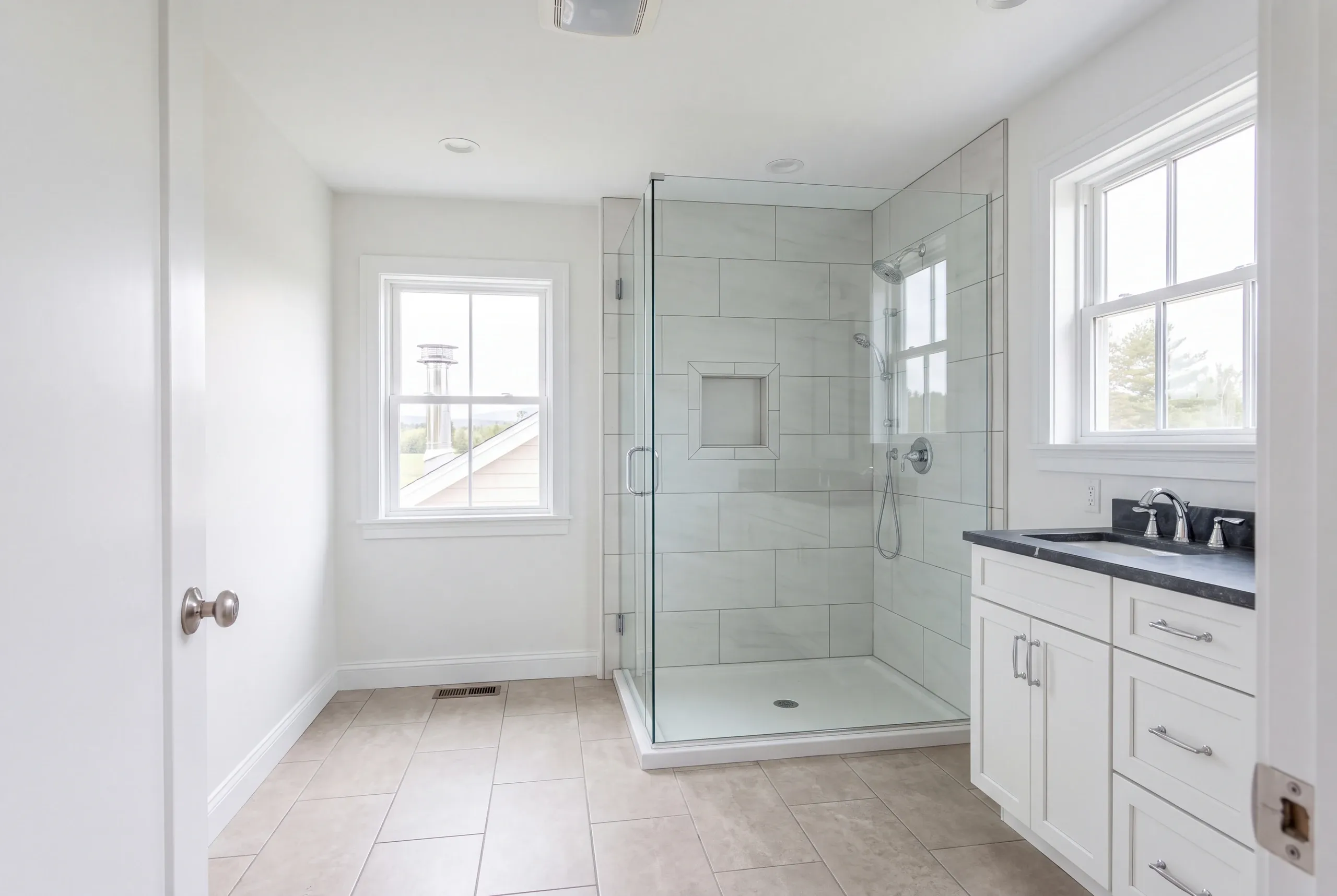 Bathroom remodeler Winslow Maine: glass corner shower with large white tile, a white vanity with a black top and two windows