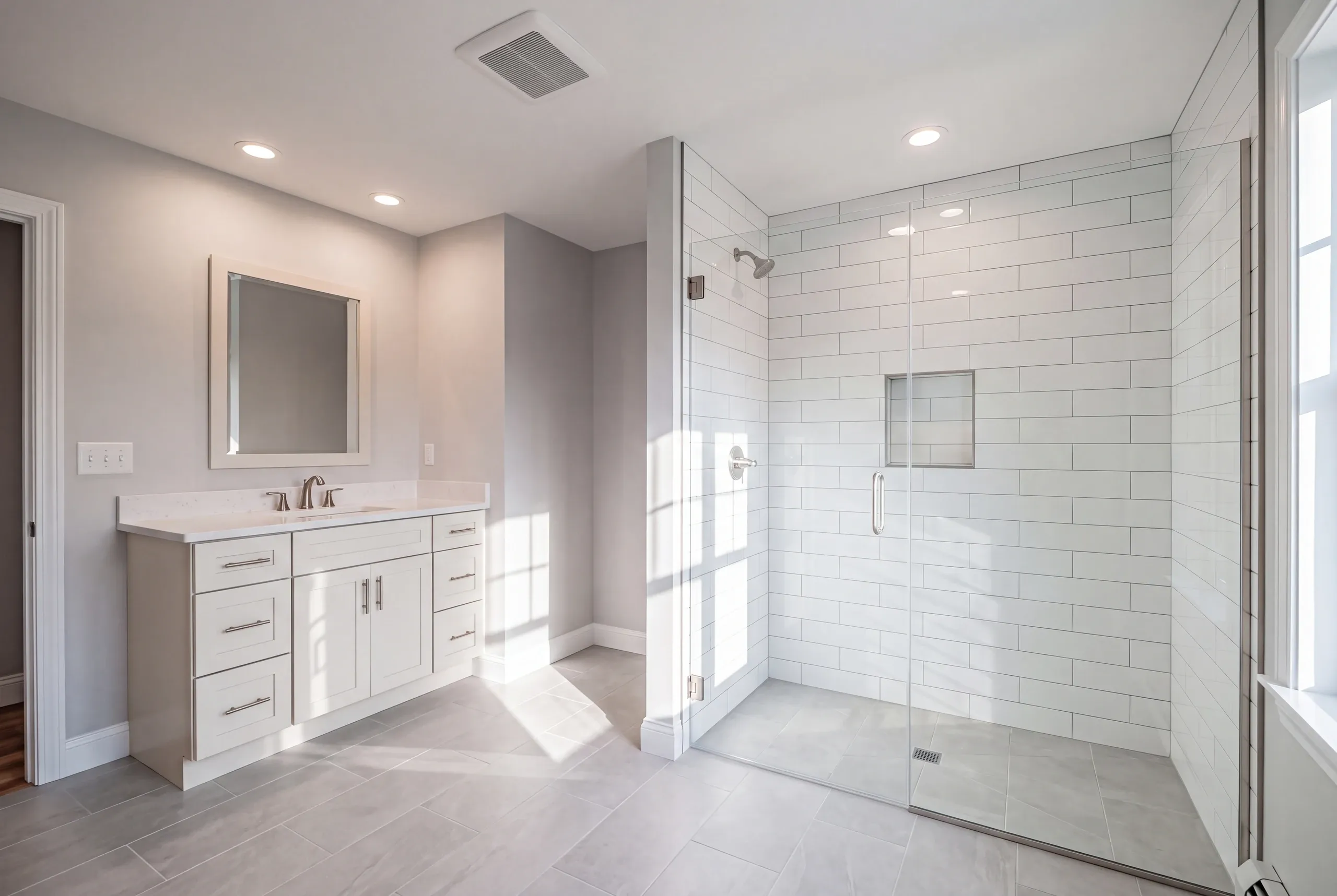 Bathroom renovation contractor Winslow Maine: curbless glass subway tile shower beside a white vanity in a sunlit gray bathroom