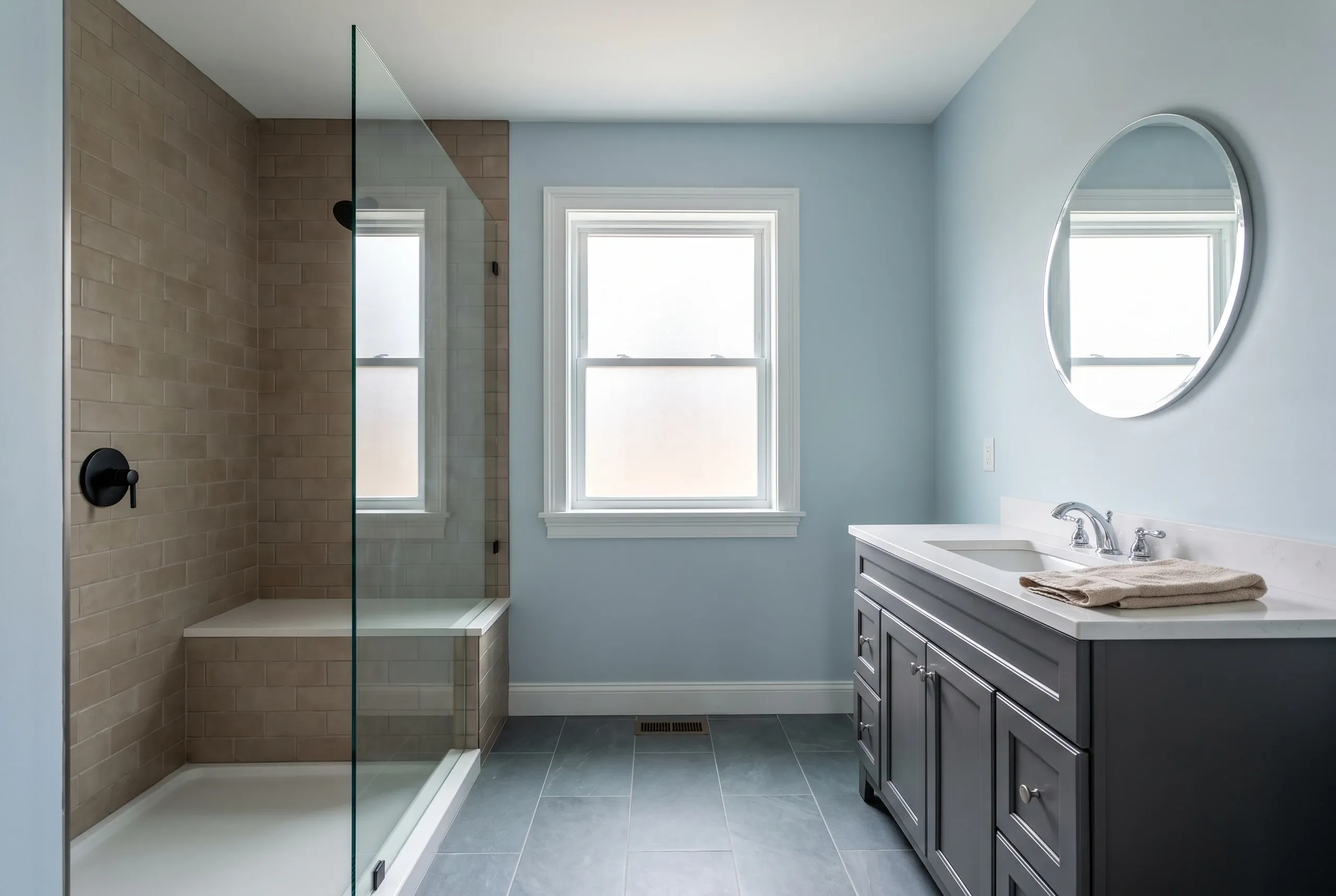 Cost to remodel a bathroom in Midcoast Maine: remodeled bath with a tan tiled walk-in shower, gray vanity with white quartz top and round mirror