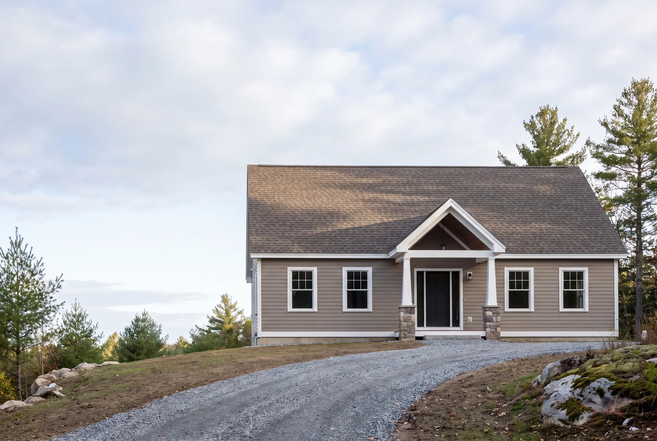New home builder Augusta Maine: single-story taupe ranch with a gabled entry on stone piers at the top of a gravel drive