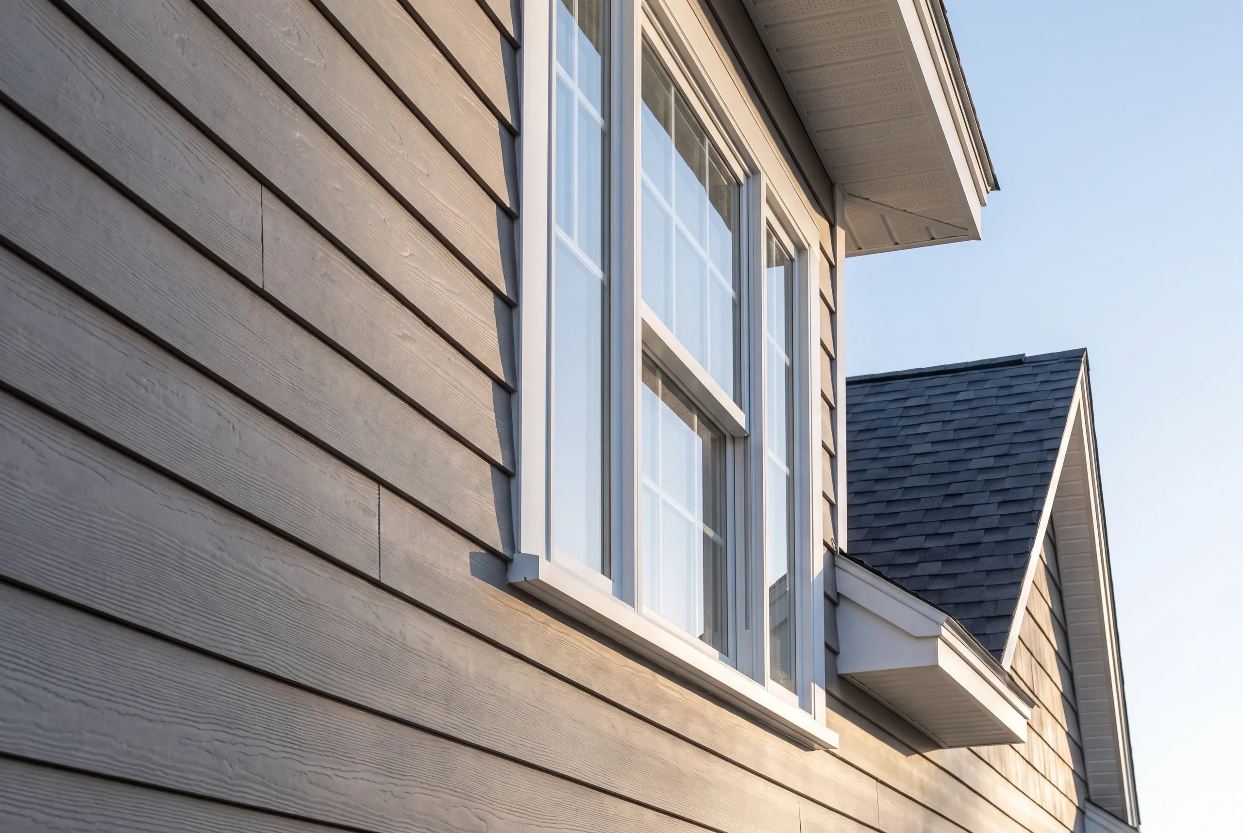 New home builder Bucksport Maine: upward view of taupe lap siding, tall double-hung windows and a roof edge against blue sky