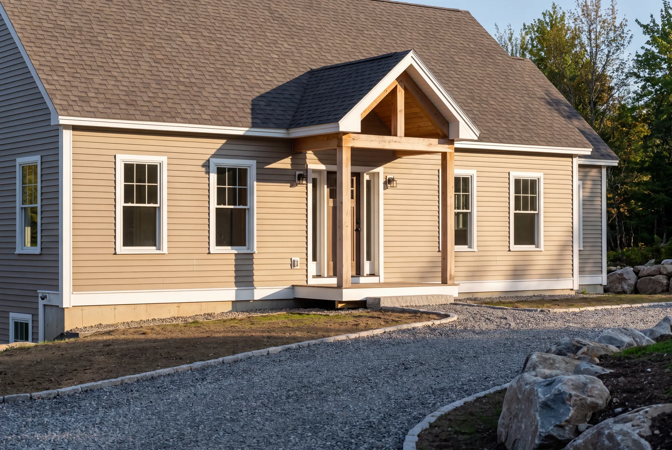 Custom home builder Camden Maine: front view of a tan cape with a timber gable entry and boulders at the edge of the drive