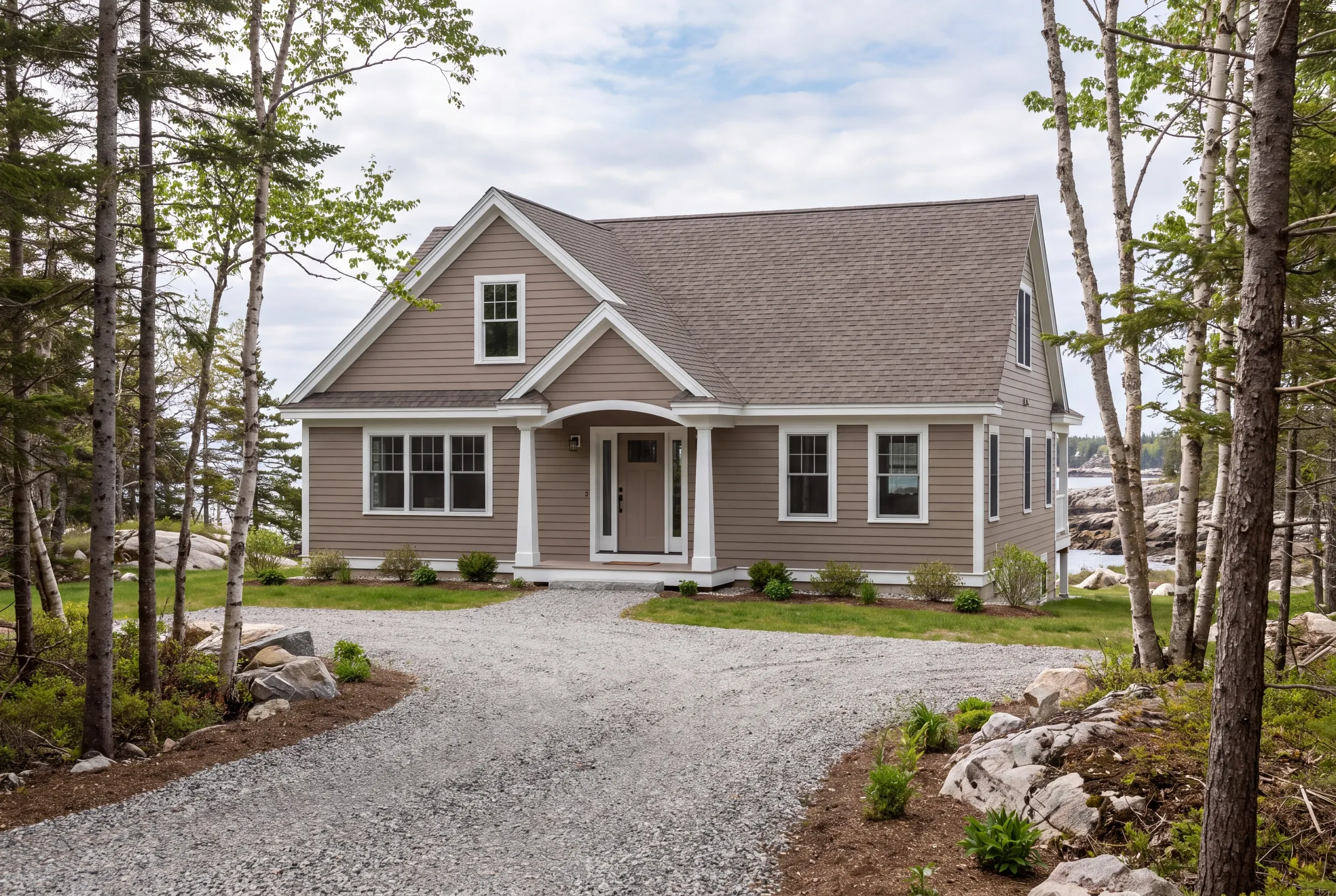Residential home builder Rockport Maine: finished taupe cape seen down a curving gravel drive past pines and ledge boulders