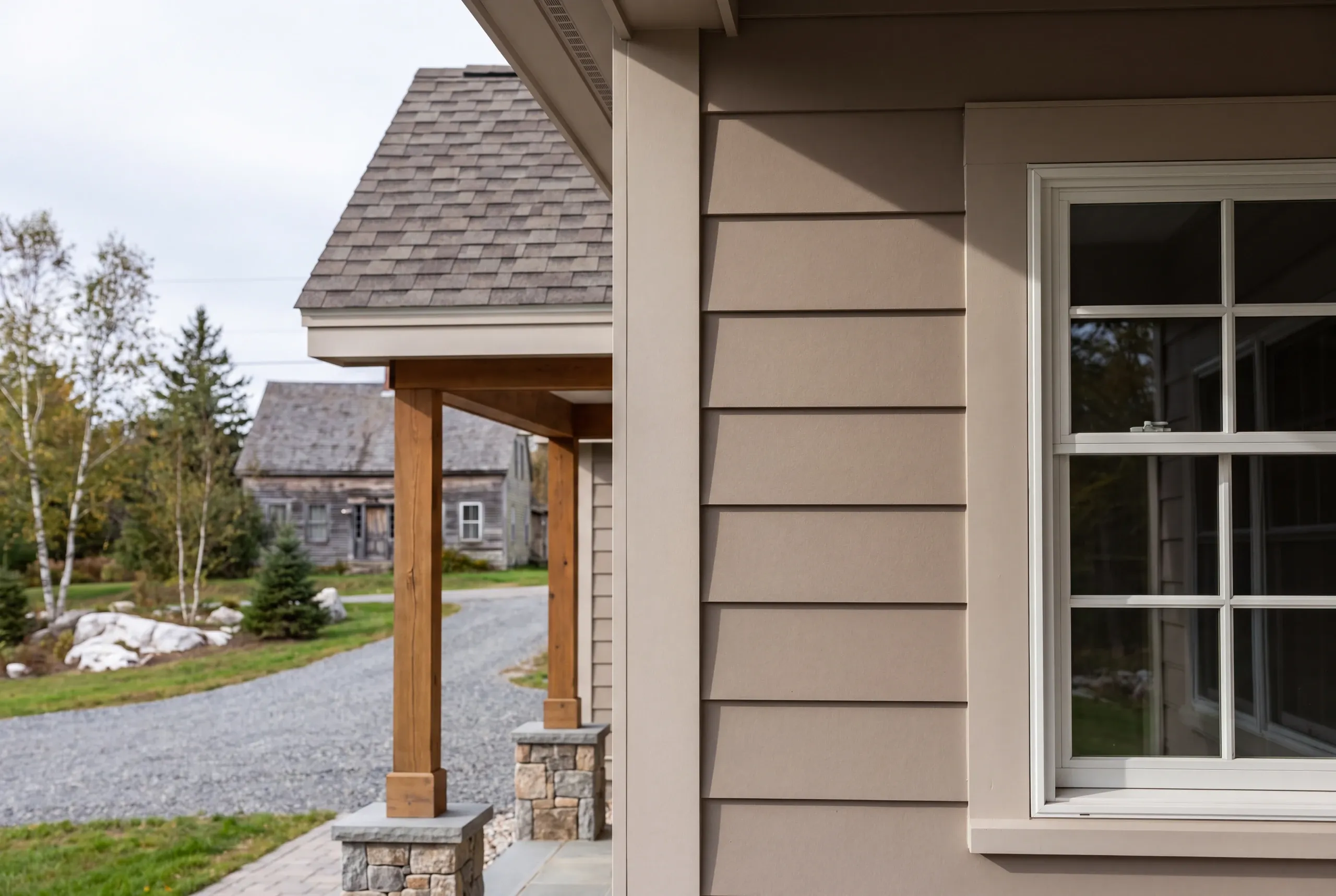 New home builder Waldoboro Maine: close view of taupe siding and timber porch posts on stone piers, an old gray farmhouse beyond