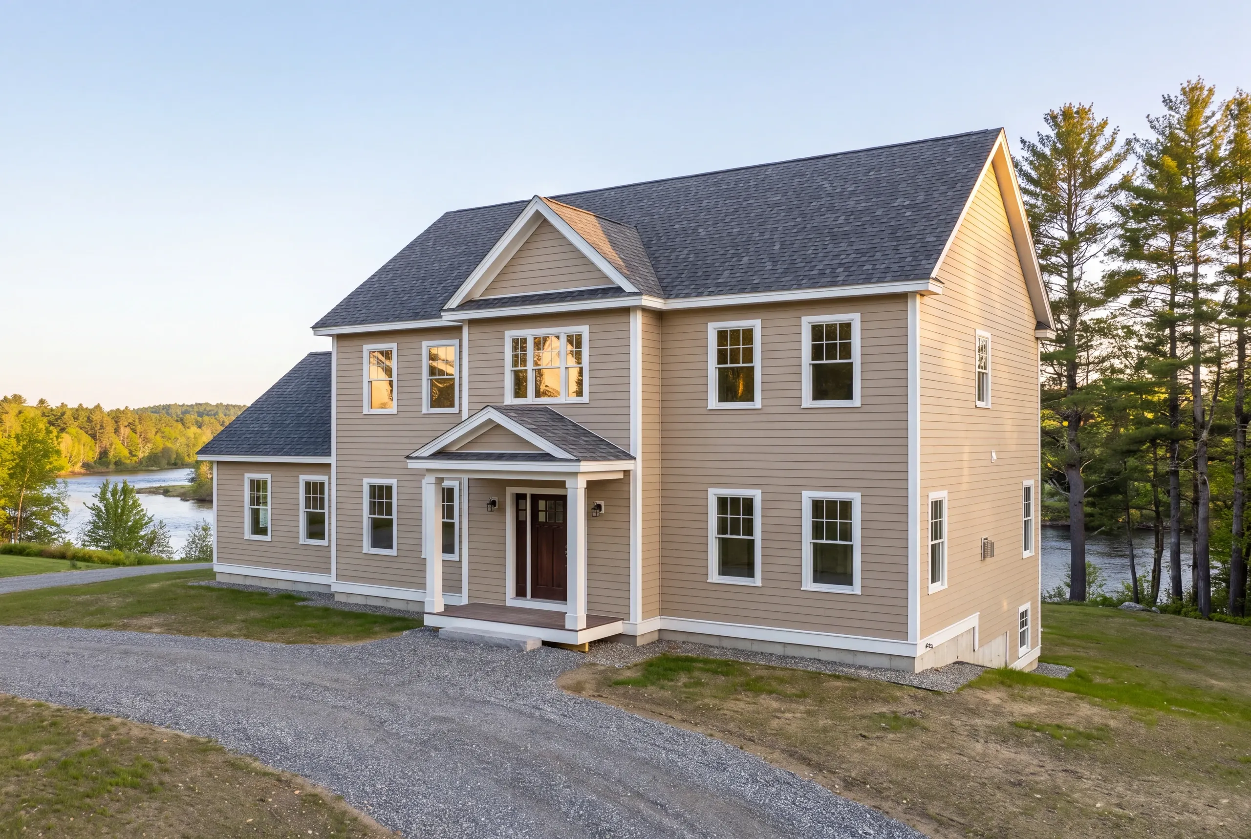 New home builder Winslow Maine: two-story tan colonial with gabled entry portico, gravel drive and water beyond