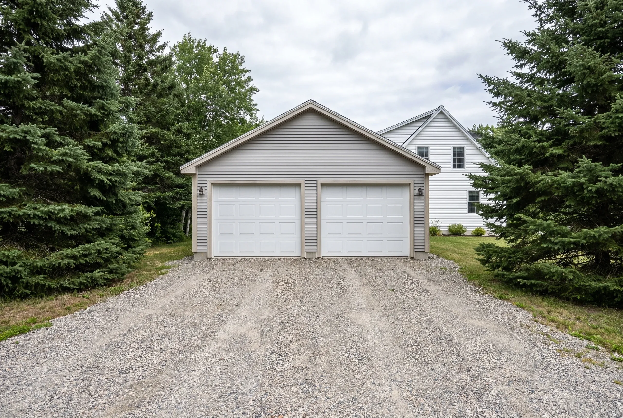 Detached garage builder Bath Maine: taupe two-bay garage with white doors on a long gravel drive beside a white farmhouse