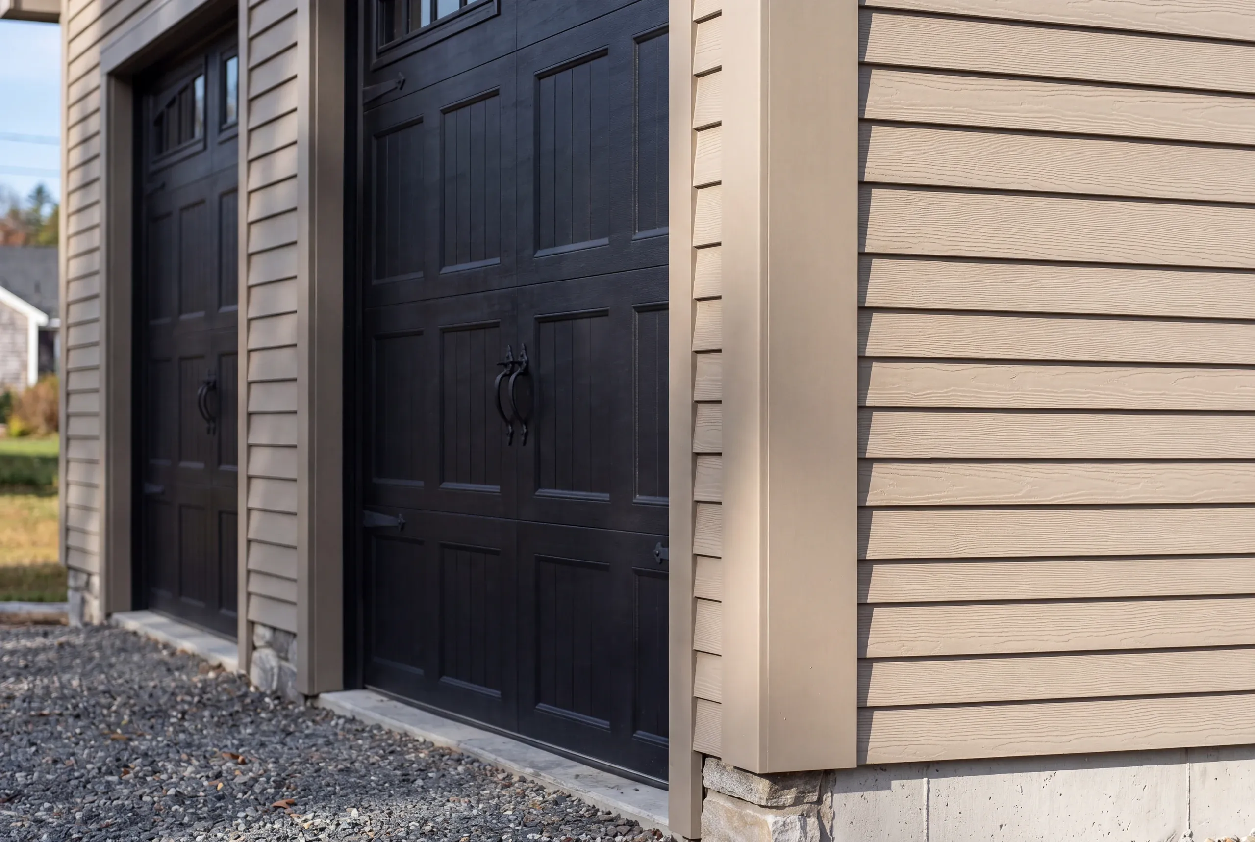 Custom garage builder Rockland Maine: close view of black carriage doors, tan lap siding and a stone-faced foundation corner