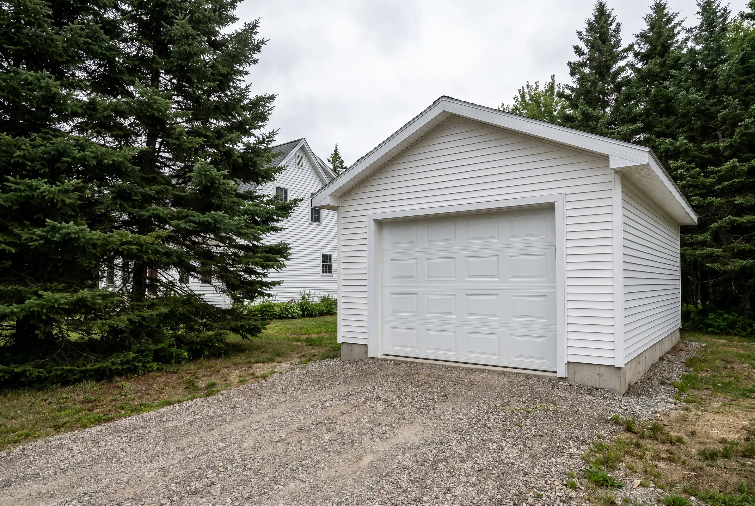Garage builder Rockport Maine: detached one-car white vinyl-sided garage with white sectional door on a gravel drive beside spruce trees
