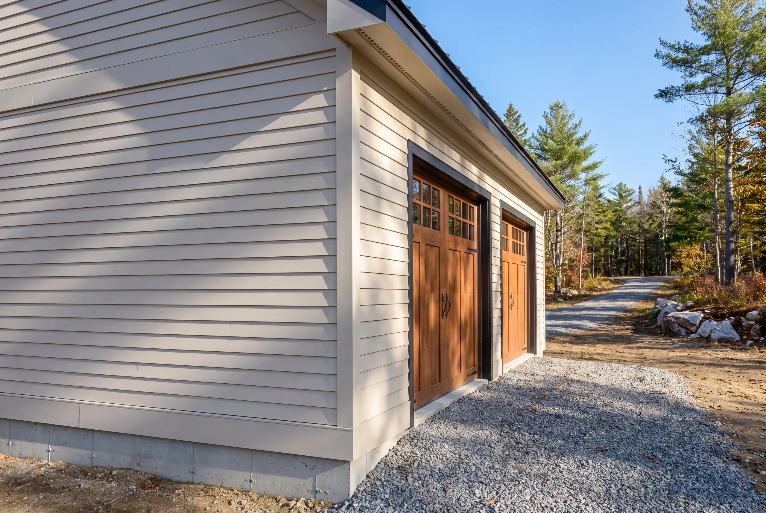 Custom garage builder Searsmont Maine: close corner of a gray lap-sided garage with wood-tone carriage doors and a drive into the woods