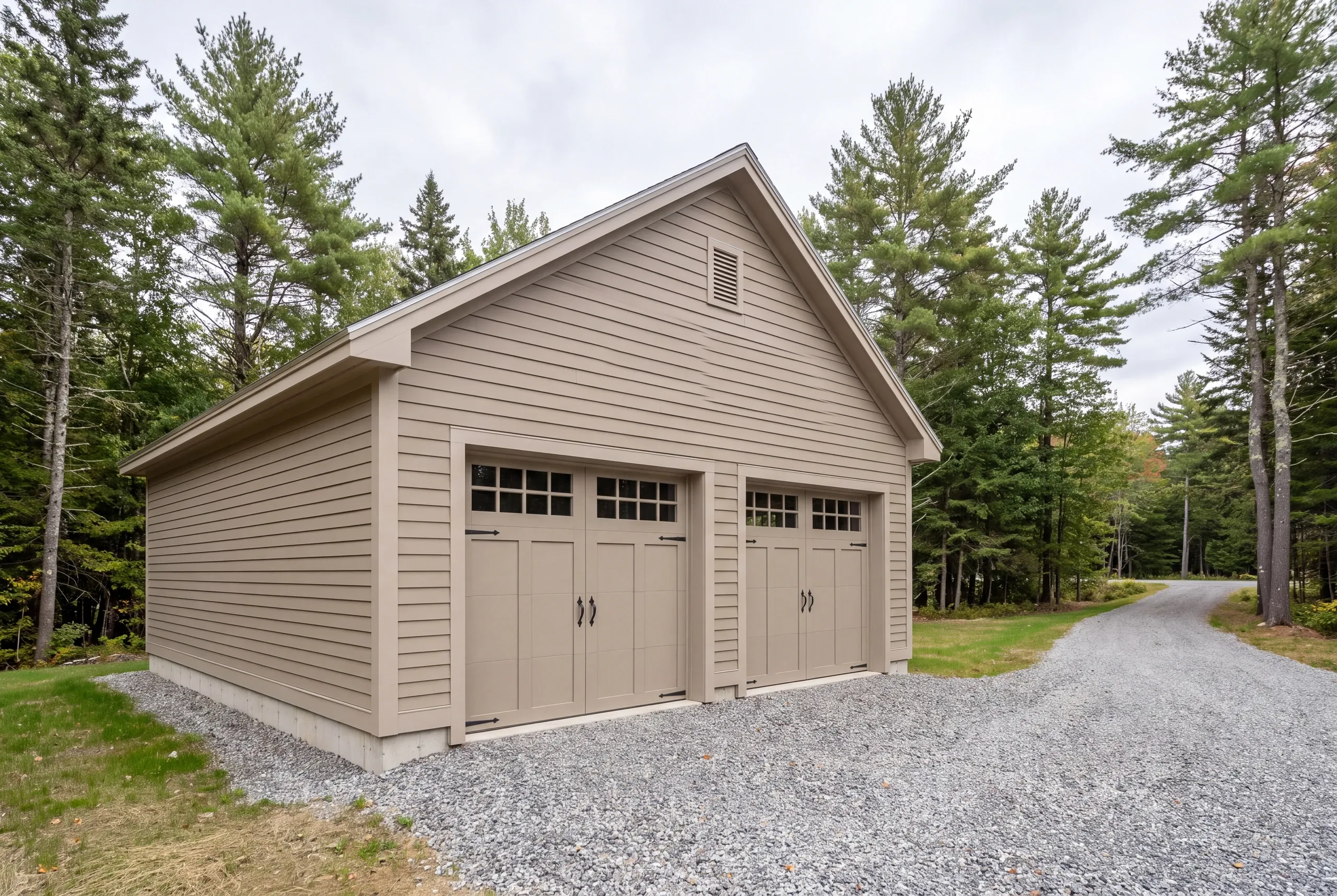 Garage builder in Warren, ME: tan two-bay garage with a gable vent and windowed doors at the end of a drive in the pines