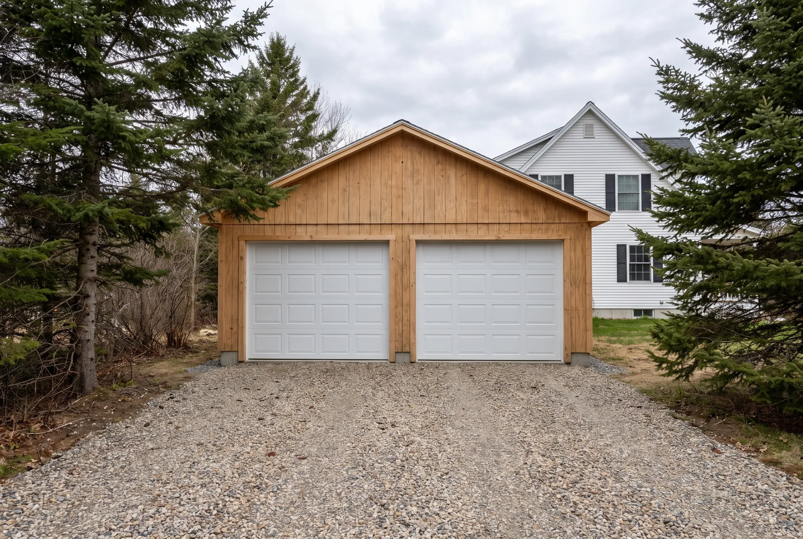 Custom garage builder Windsor Maine: new pine board-and-batten two-bay detached garage with white doors on raw gravel between evergreens