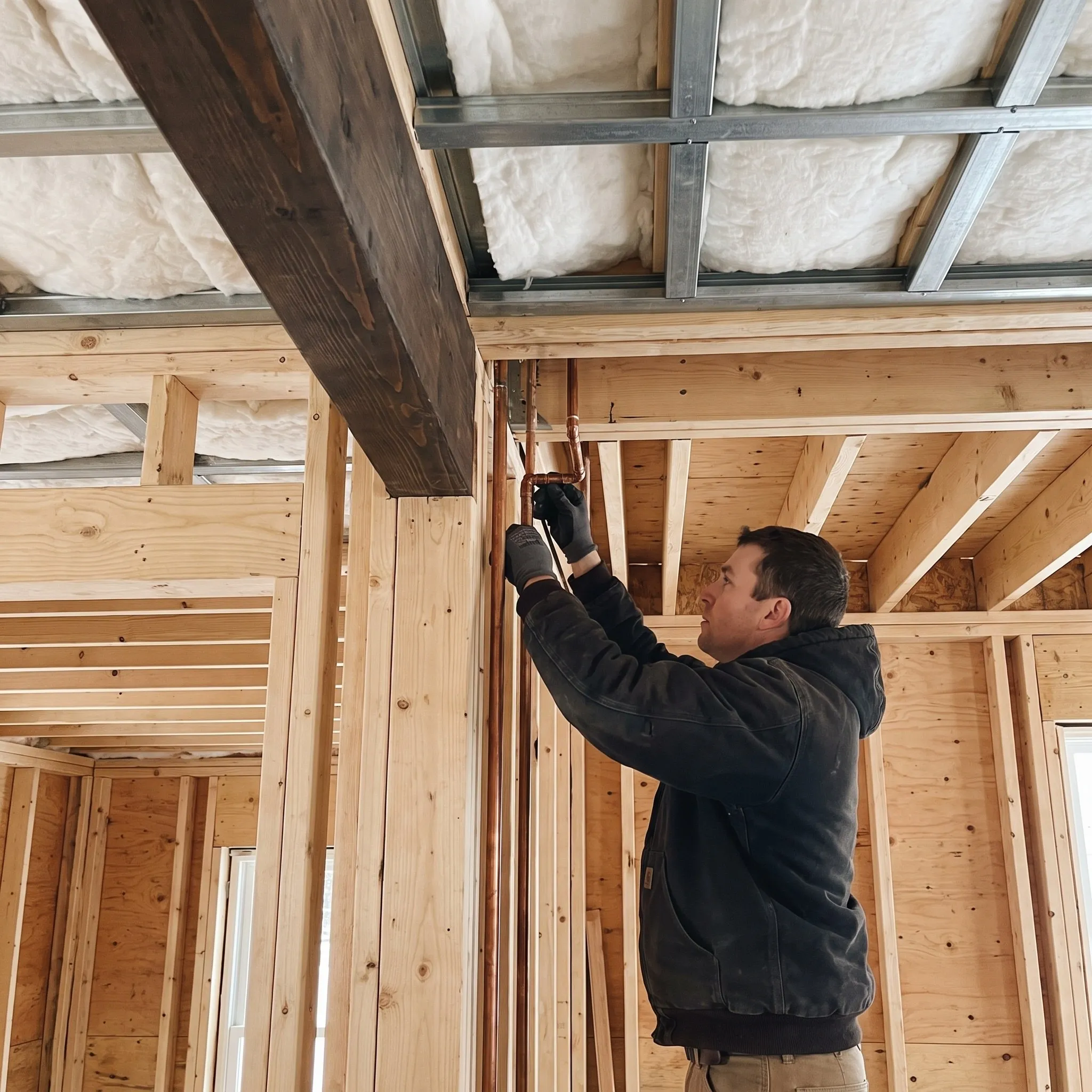 Residential general contractor Gardiner Maine: worker fitting copper pipe by a dark wood beam under an insulated steel-channel ceiling