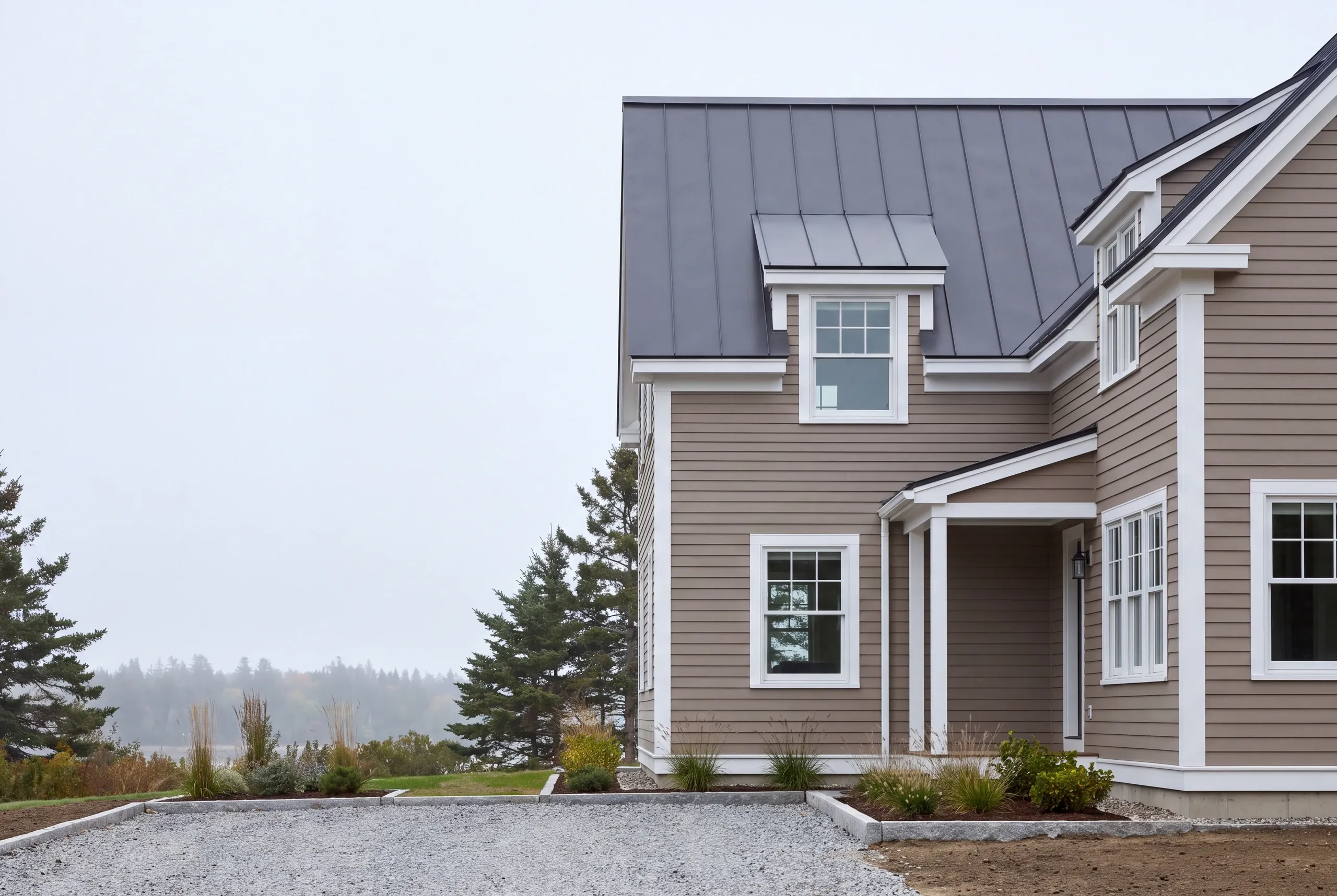 Construction company Lincolnville Maine: taupe two-story house with standing seam roof and dormers above a foggy shore