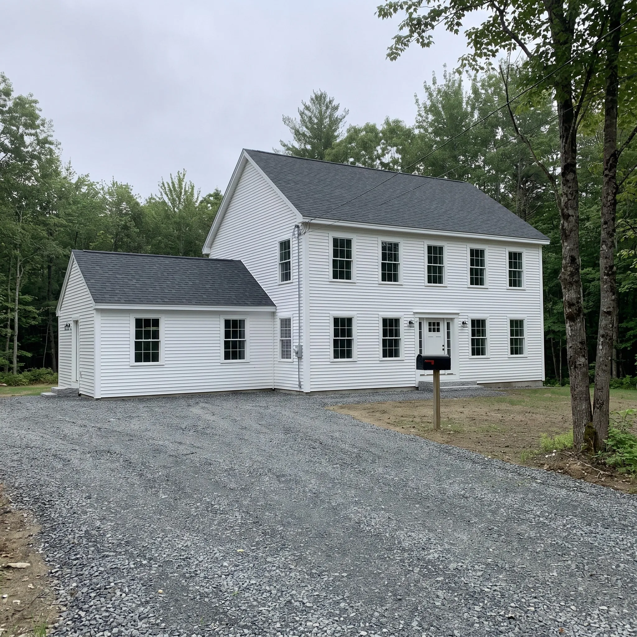 Residential general contractor Winslow Maine: white colonial with symmetric windows, a side ell and a gravel drive at the edge of the woods.