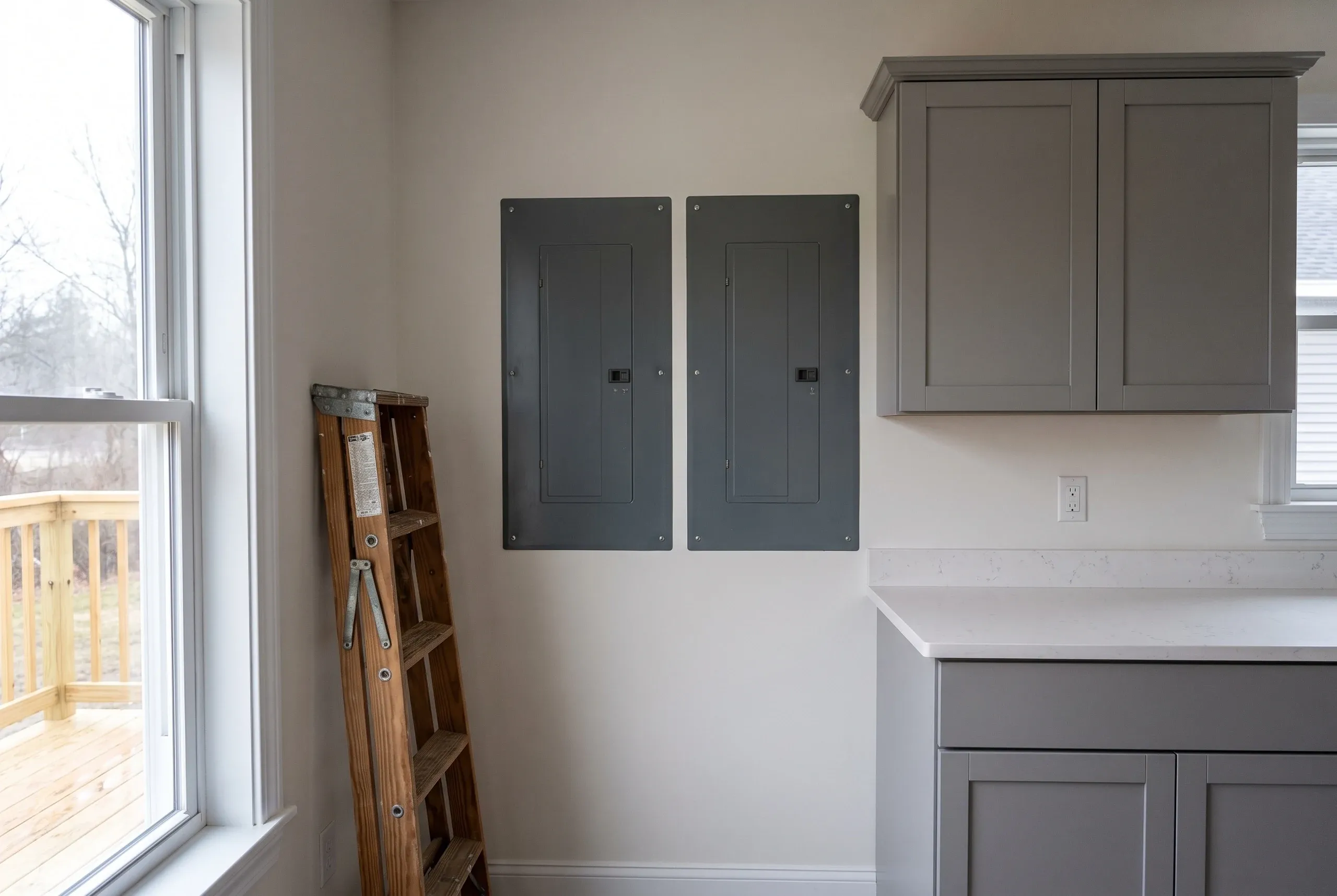 Kitchen renovation contractor Augusta Maine: two breaker panels on a clear wall beside gray shaker cabinets and a quartz-look counter