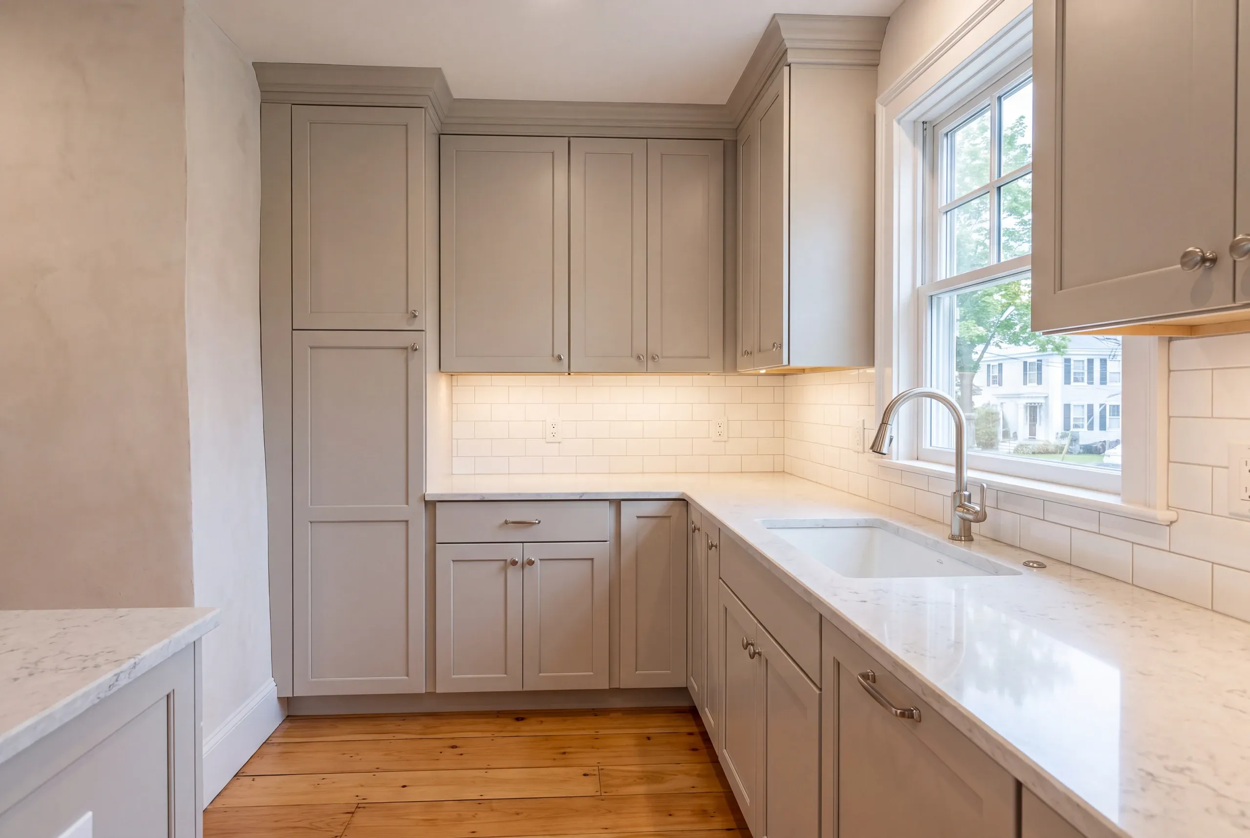 Kitchen remodeler Bath Maine: tall pantry cabinet beside a wavy plaster wall, quartz-look counter and white sink under a window
