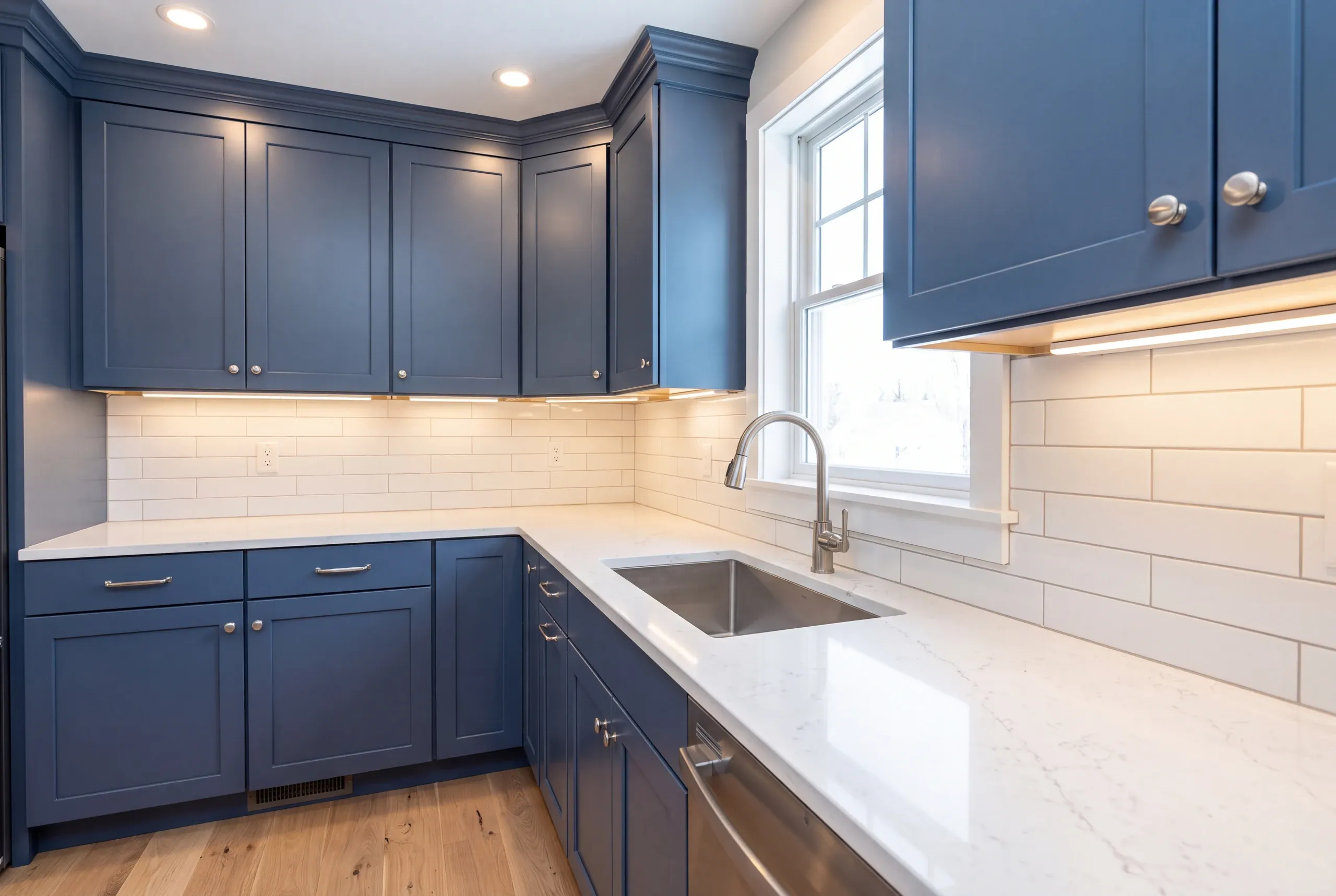 Kitchen renovation contractor Belfast Maine: blue shaker cabinets wrapping a corner with long white tile and an undermount sink under a window