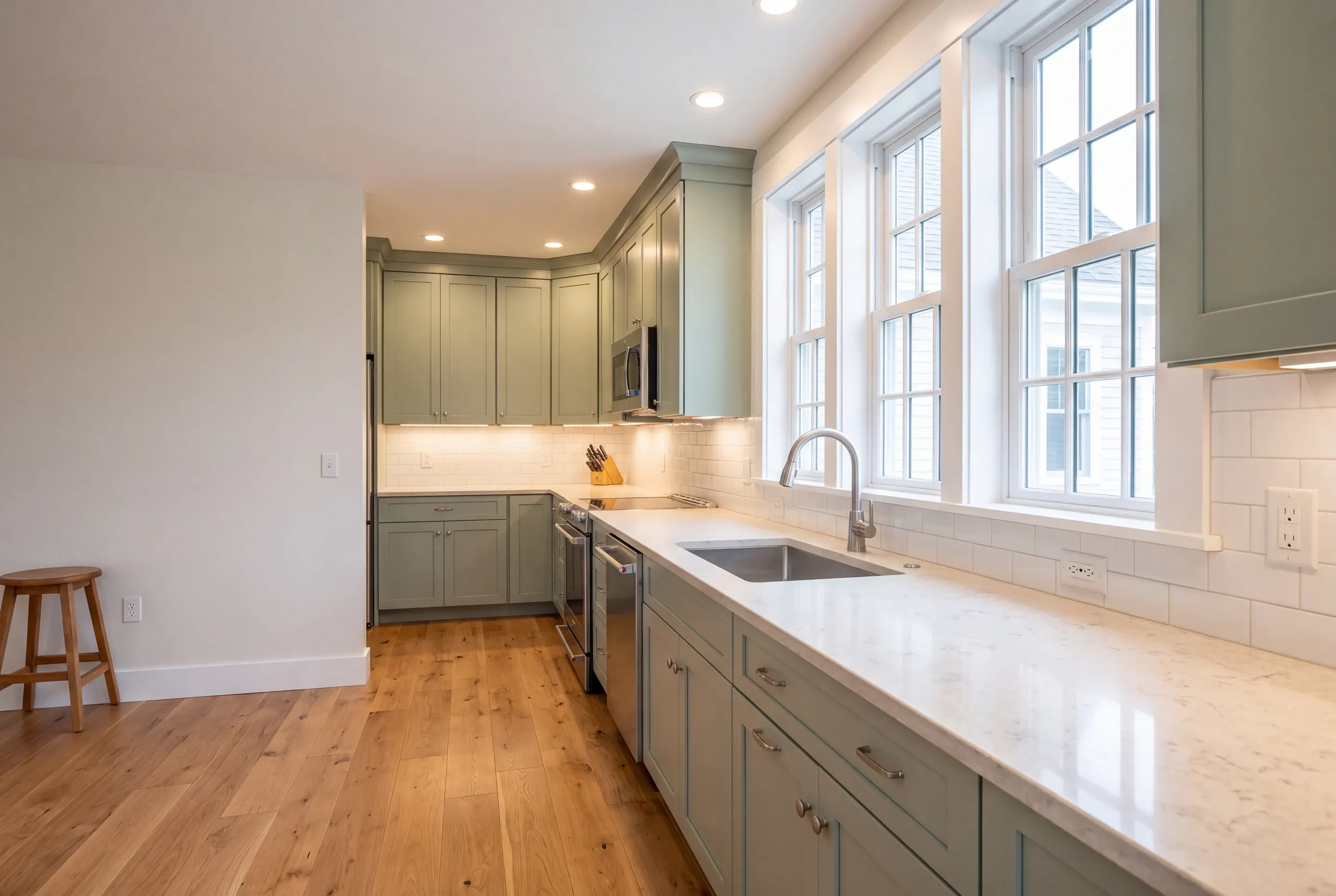 Kitchen remodeling contractor Midcoast Maine: sage green shaker kitchen with tall windows, a wood stool and wide oak flooring