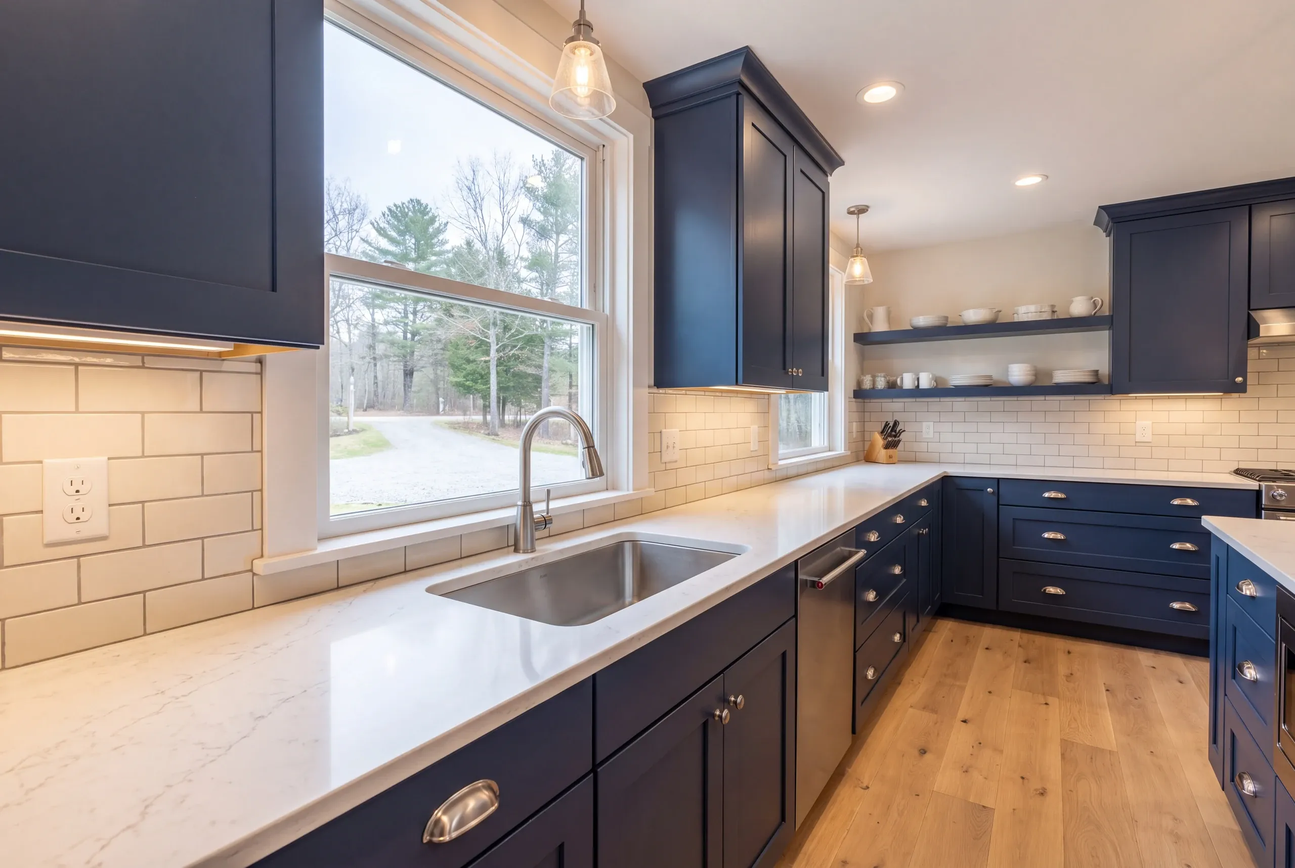 Kitchen remodeler Damariscotta Maine: navy shaker kitchen with open shelves, glass pendants and a big sink window facing a gravel drive