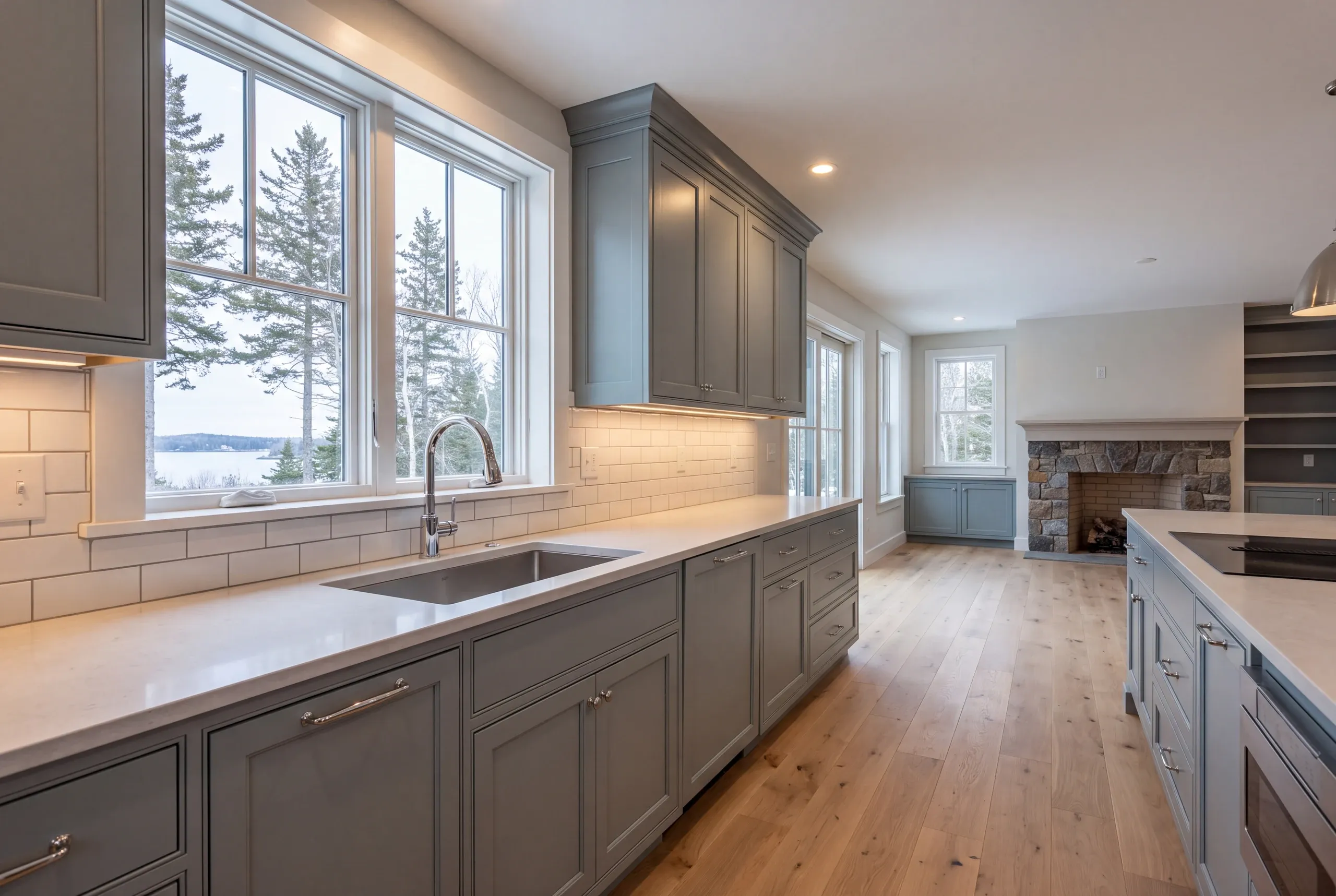 Kitchen remodeling in Lincolnville, ME: slate blue inset kitchen with a lake view sink window, cooktop island and stone fireplace beyond