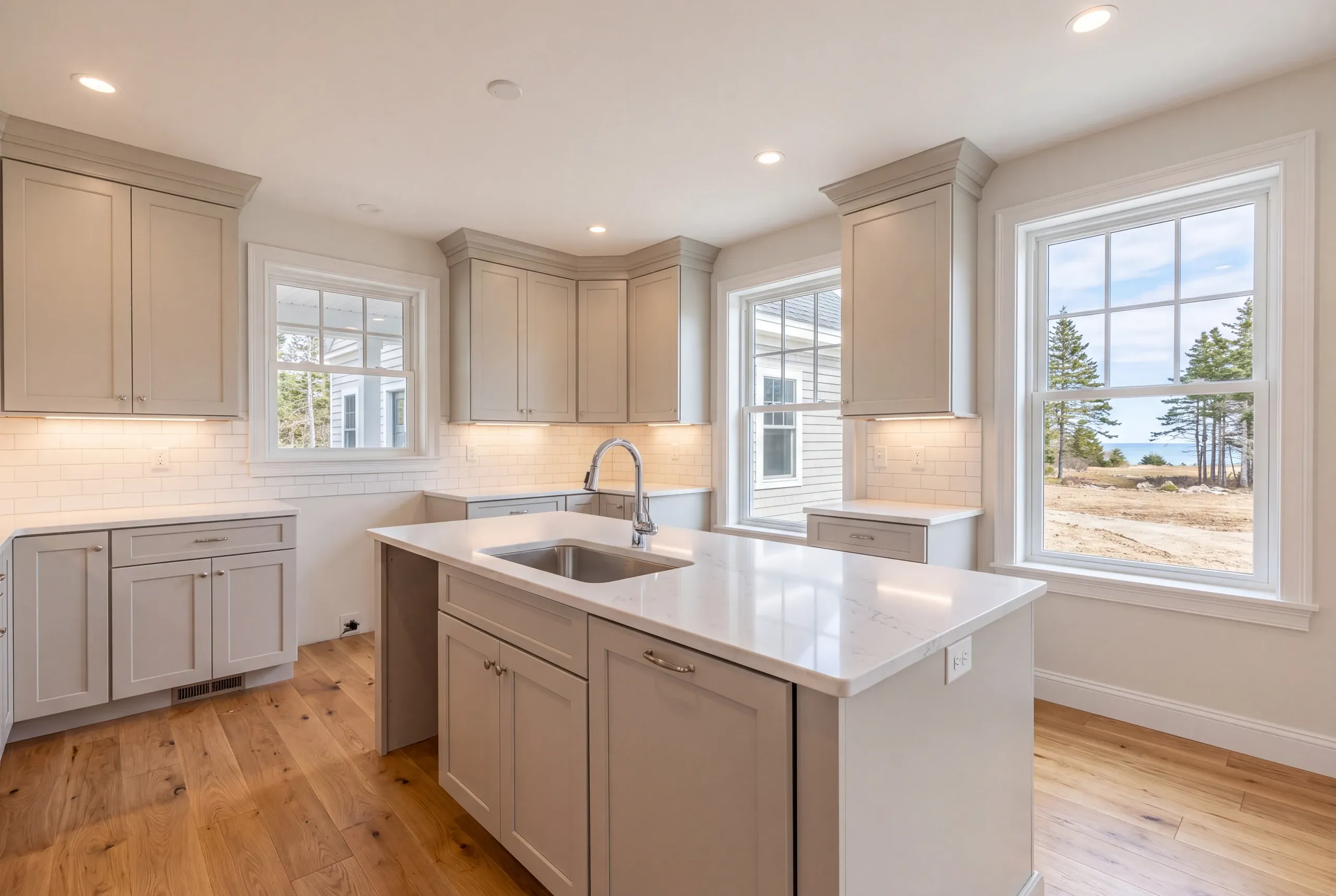 Kitchen remodeler Lincolnville Maine: greige shaker kitchen with a sink island under windows facing the ocean across a graded yard