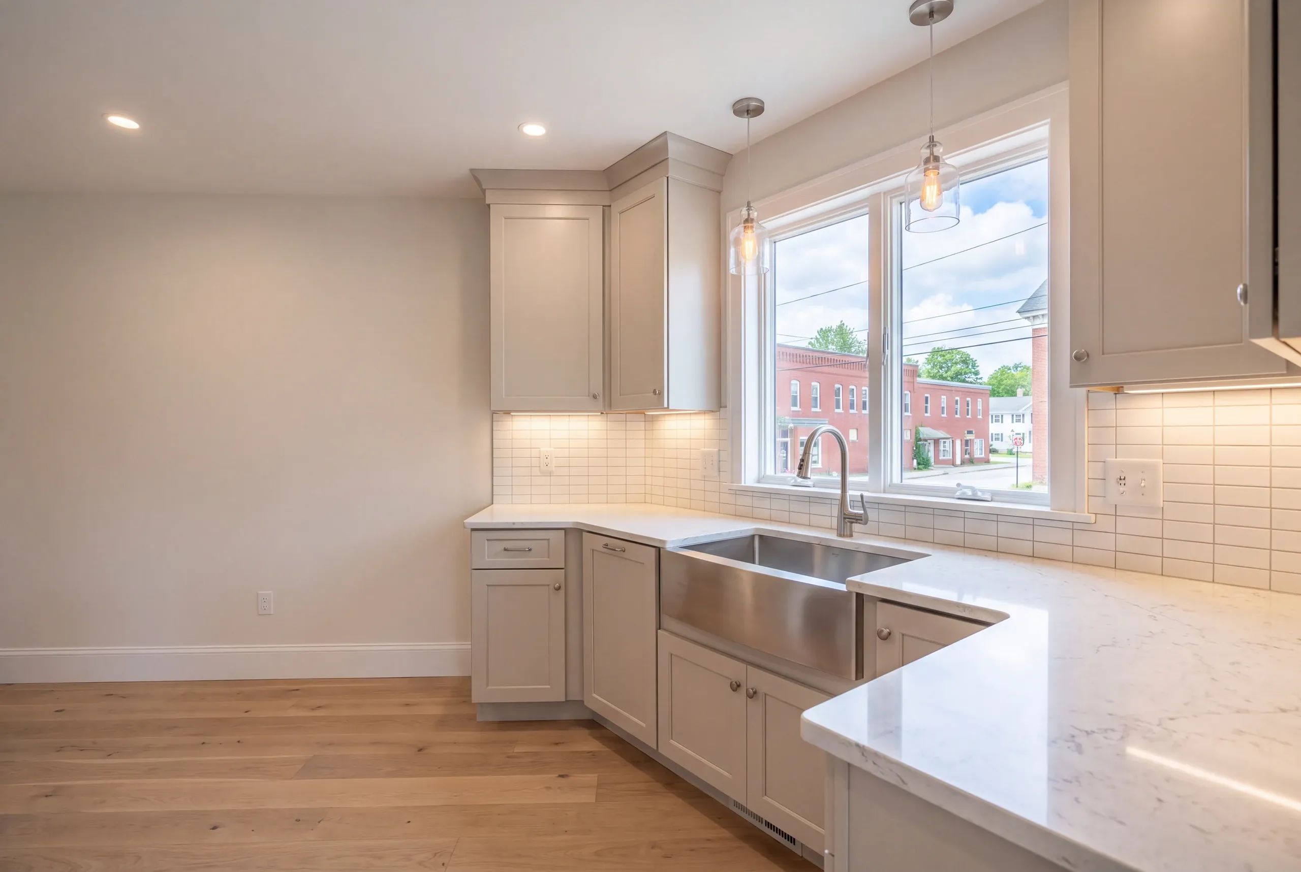 Kitchen remodeler Pittsfield Maine: greige shaker corner, stainless apron sink and glass pendants at a window over brick storefronts