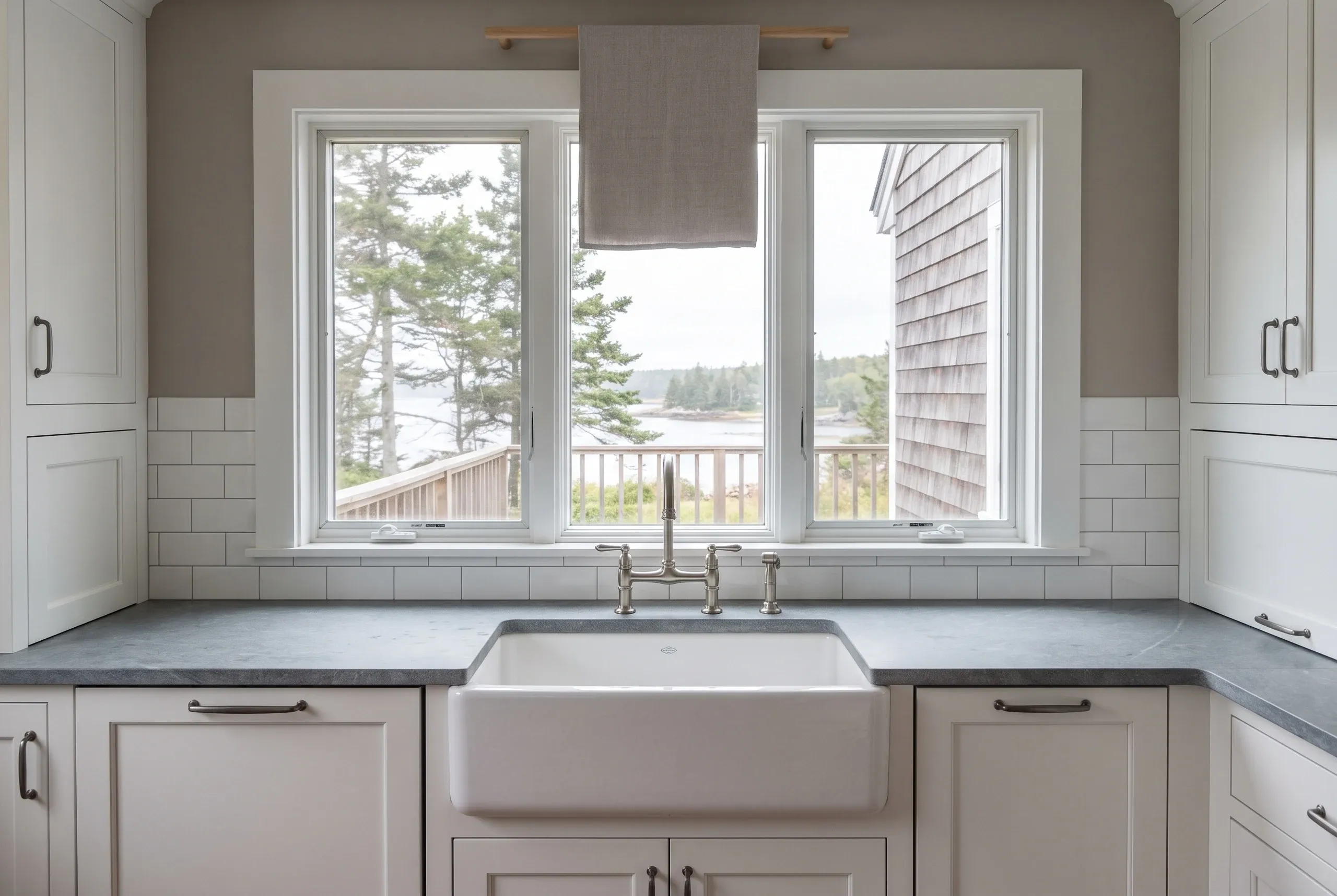 Kitchen renovation contractor Rockport Maine: fireclay apron sink and bridge faucet under a triple window facing the water, with subway tile