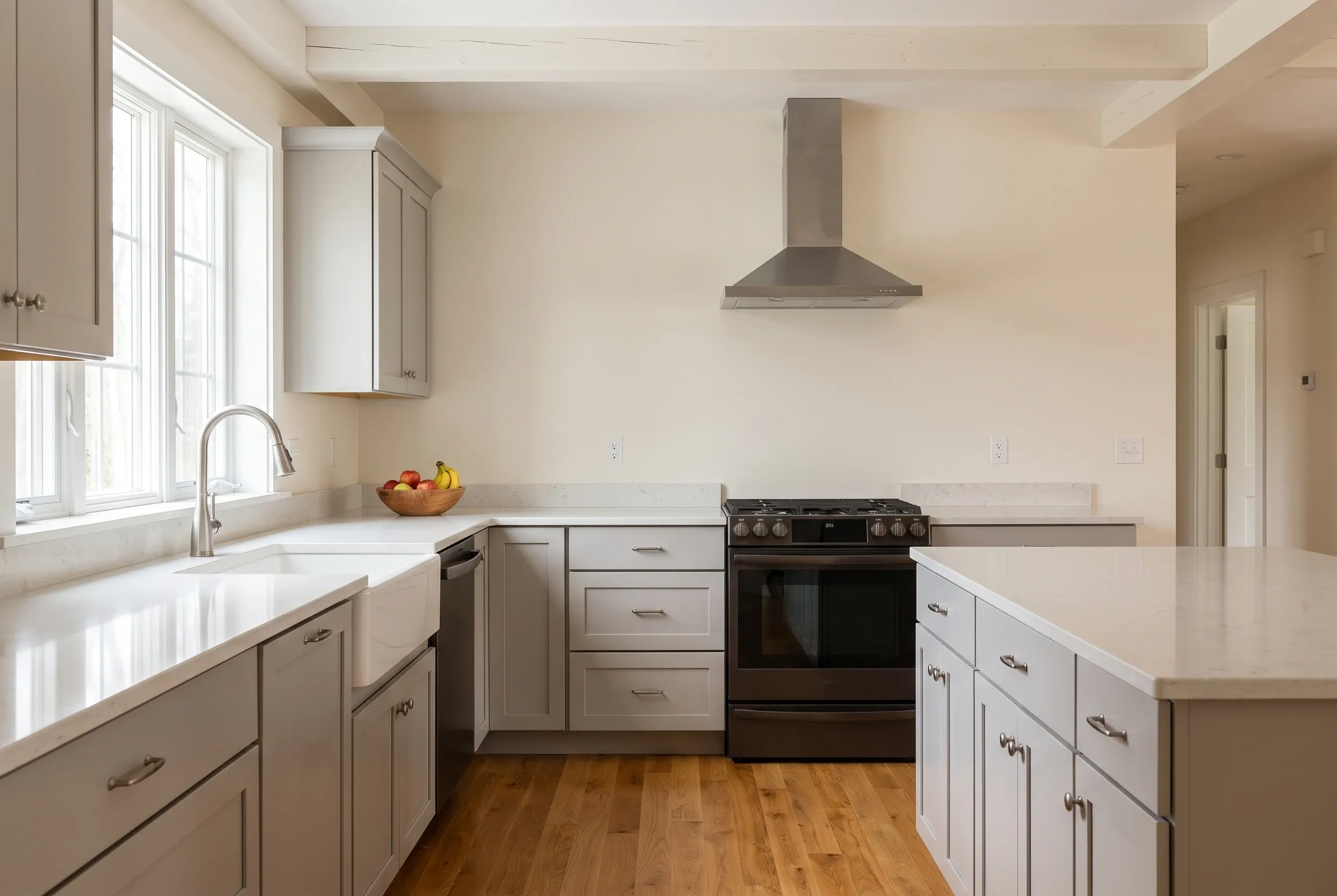Kitchen remodeling in Waldoboro, ME: light gray shaker kitchen with white quartz counters, stainless chimney hood and exposed ceiling beams