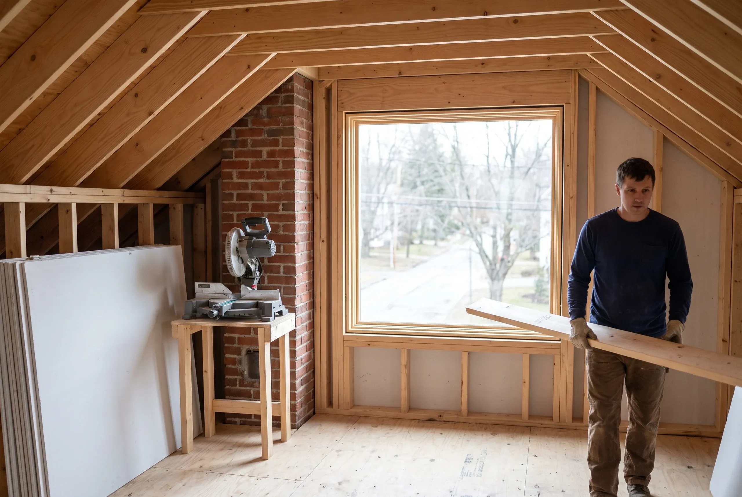 Home remodeling contractor Bath Maine: attic renovation with open framing, a large window, stacked drywall, a miter saw and brick chimney
