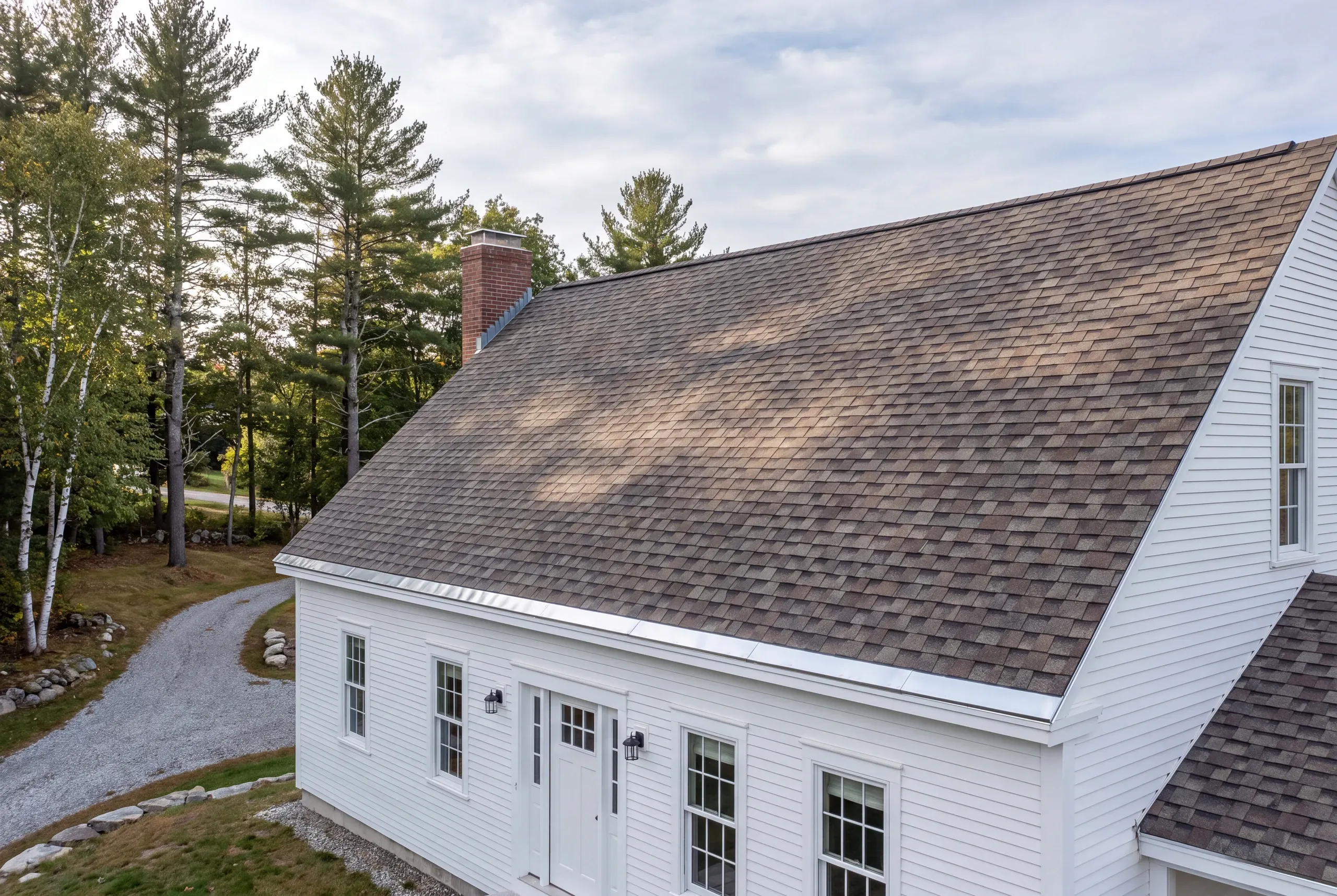 Roof replacement contractor Augusta Maine: aerial view of a white cape with a brick chimney, architectural shingles and gravel drive