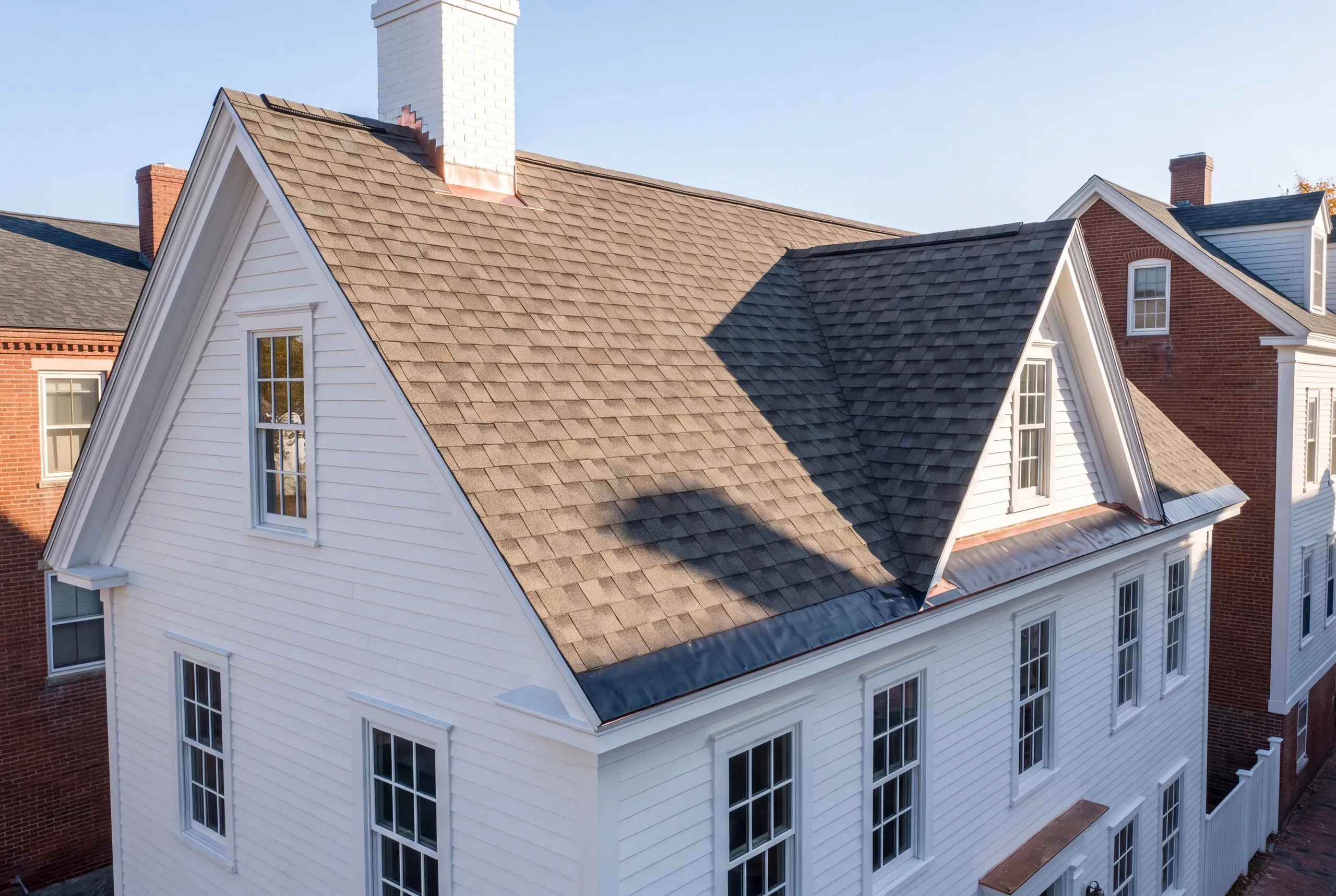 Residential roofing contractor Bath Maine: white gable-front house with a chimney and dark gray architectural shingles among brick homes