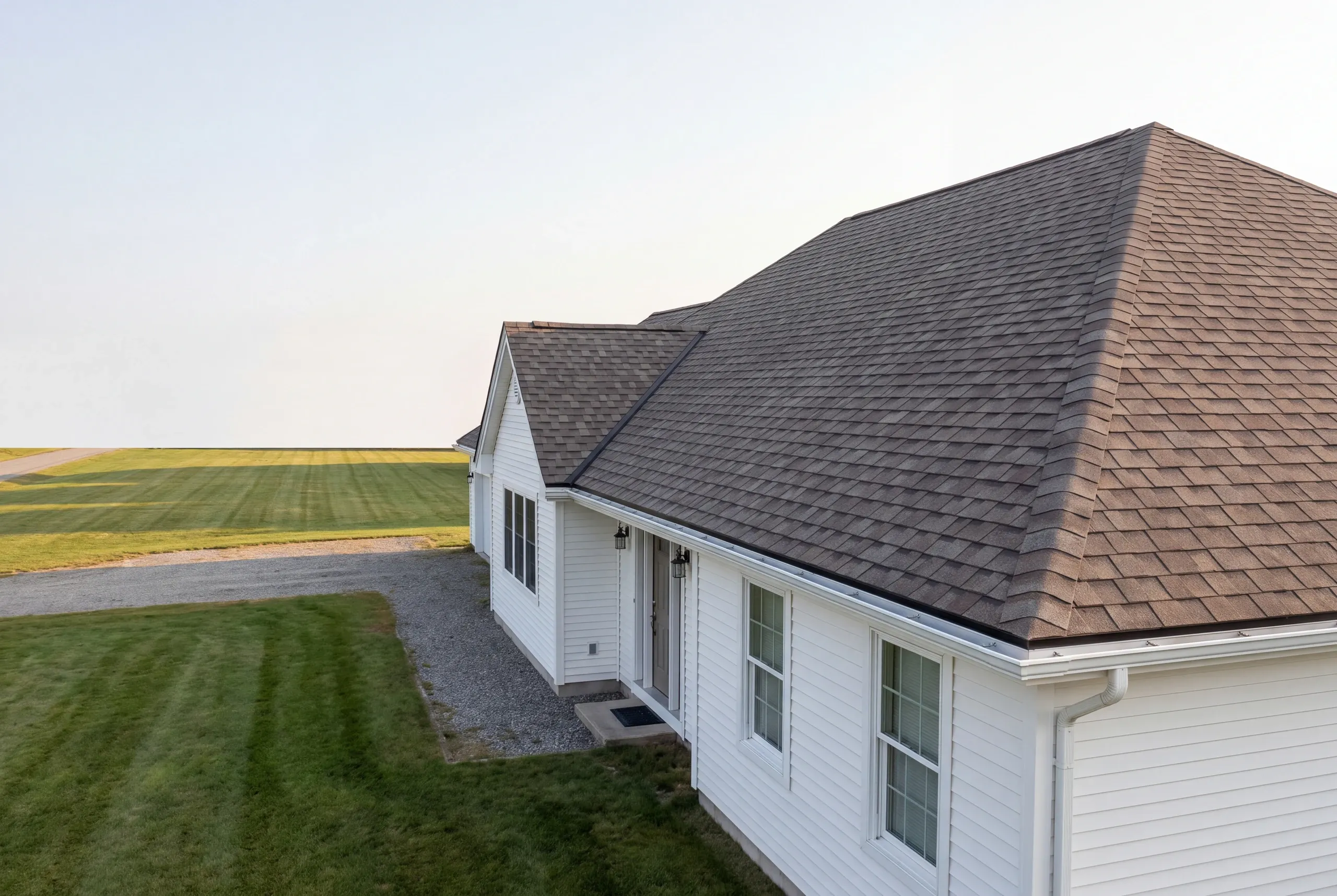 Roof replacement contractor Fairfield Maine: white ranch with a dark shingle hip-end roof beside a mowed field and gravel drive